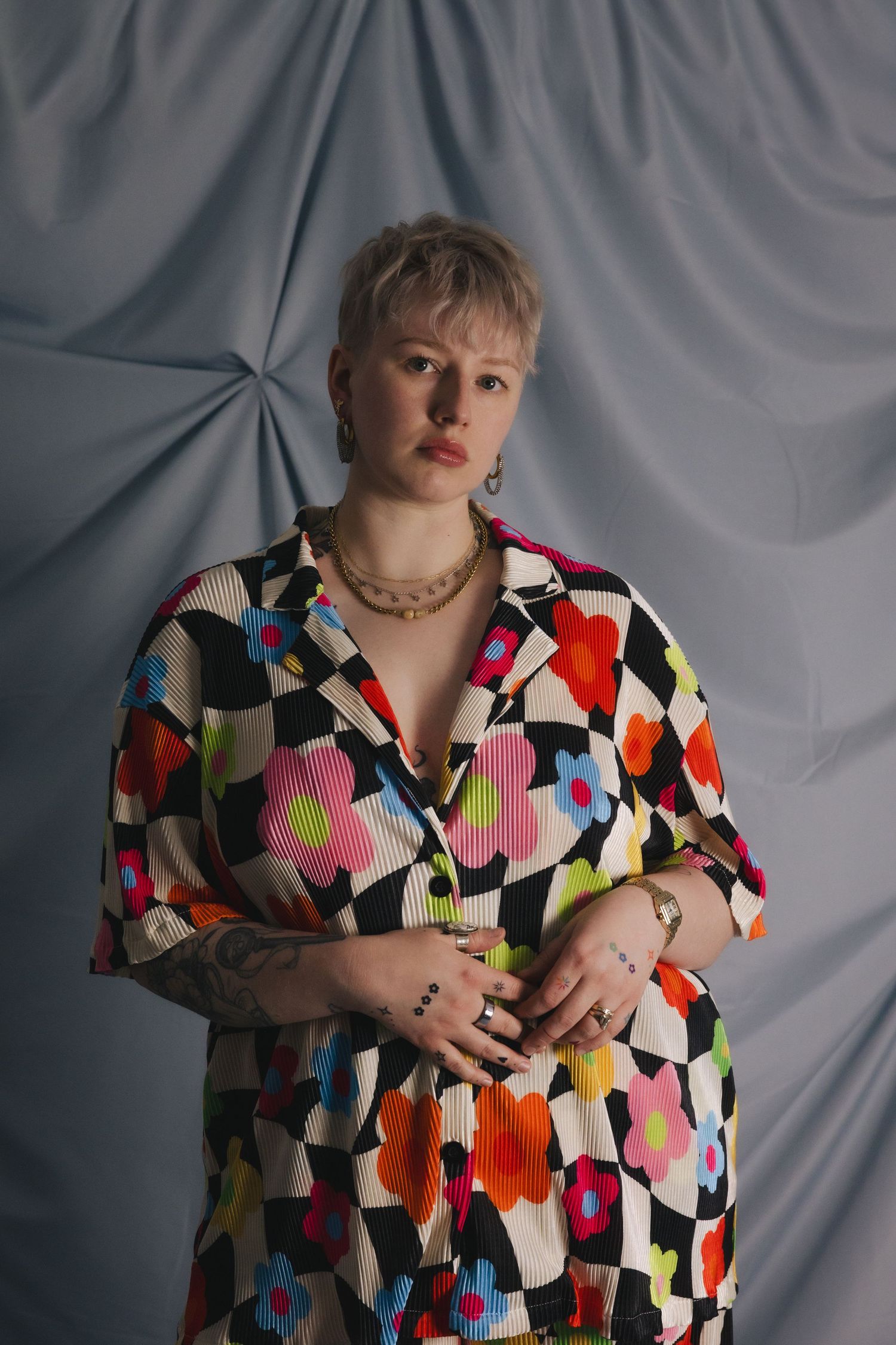 Person in colorful floral patterned shirt with short blonde hair posing against gray draped fabric backdrop.