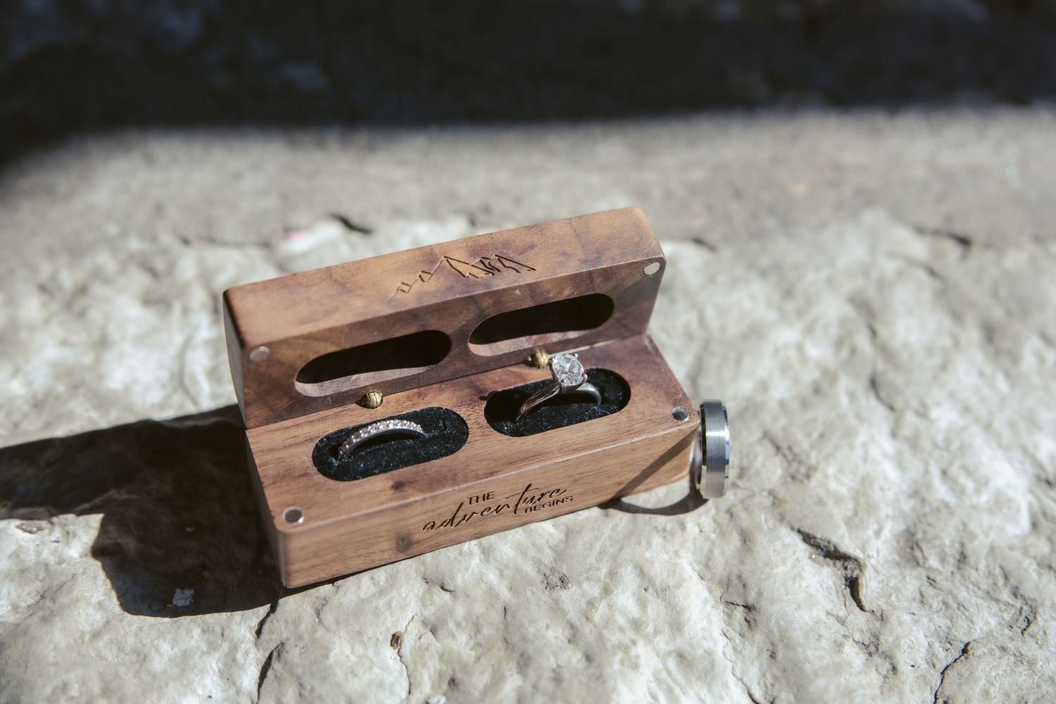 A wooden double ring box with two black wedding bands nestled inside sits on a textured stone surface in natural light.