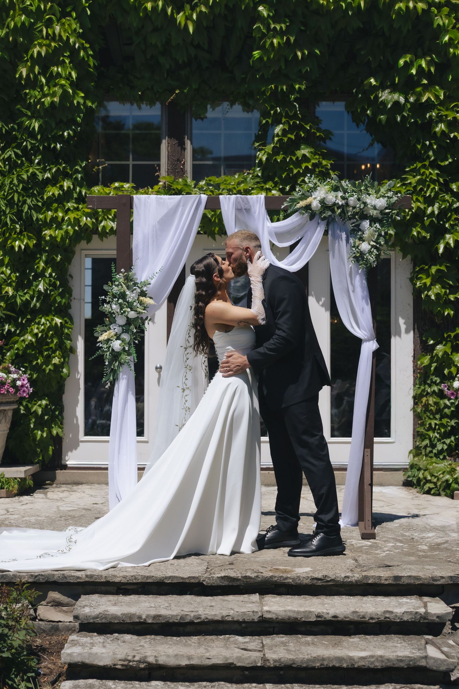 Bride and groom share first kiss at outdoor wedding altar adorned with white draping and greenery.