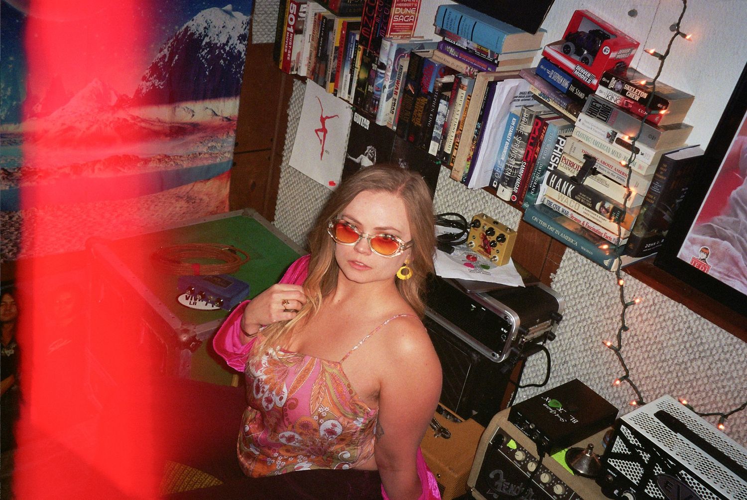 Young woman in pink floral top and sunglasses poses in a colorful, eclectic room filled with books and art.