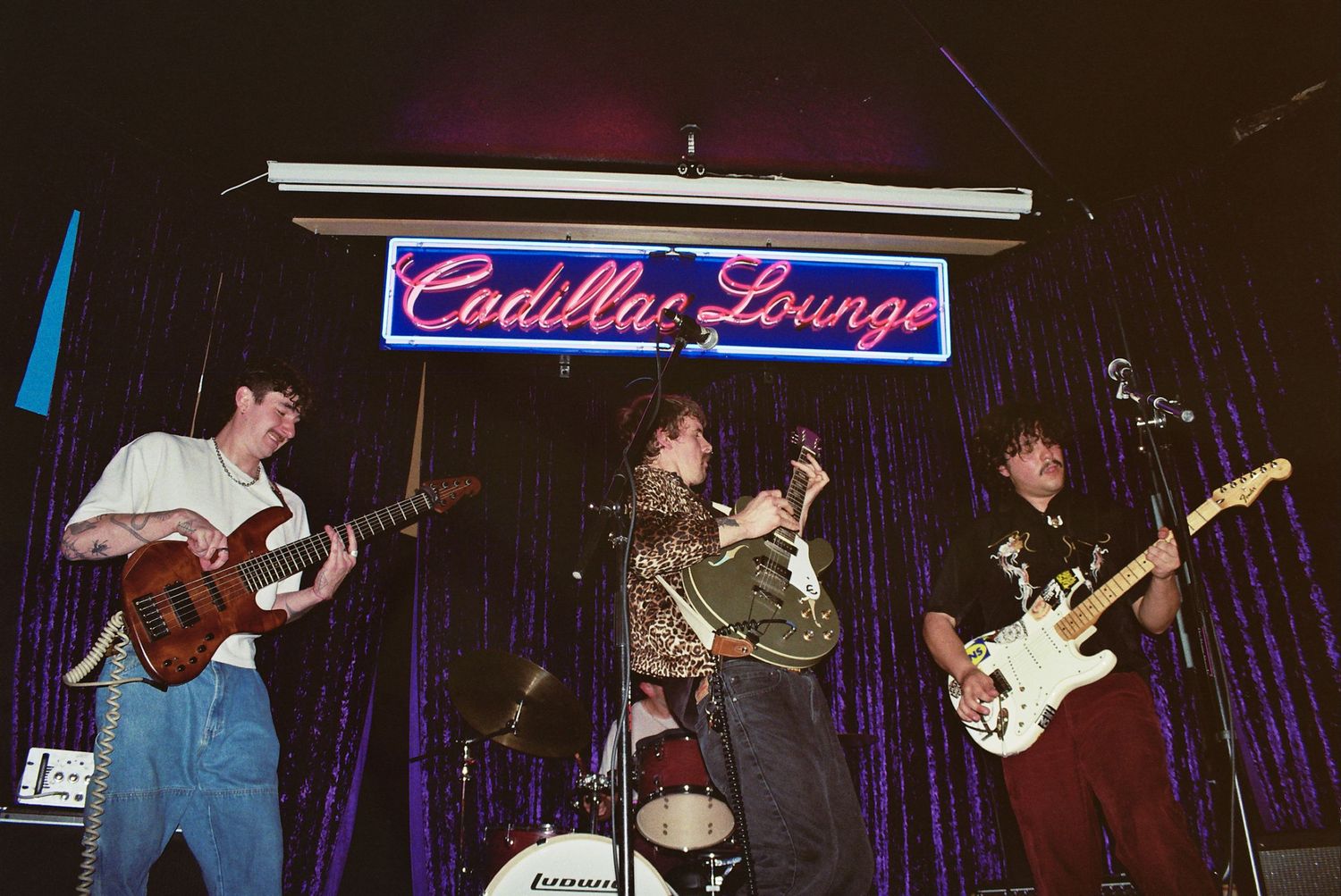 Four young musicians perform on stage at Cadillac Lounge, playing electric guitars and drums under purple neon lighting.