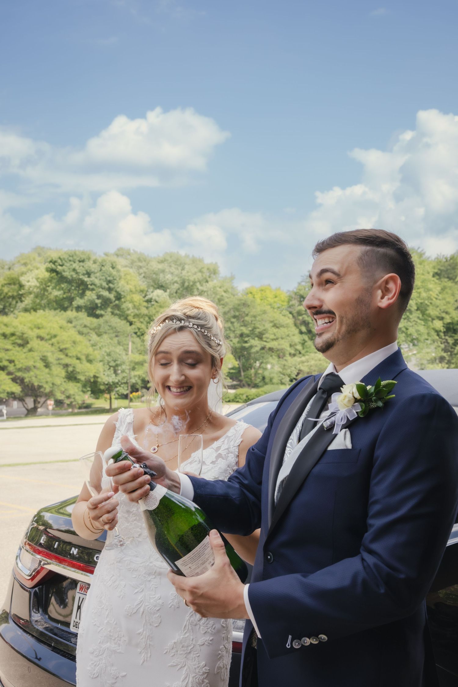 Newlyweds celebrate with champagne outdoors on a sunny day with green trees in the background.