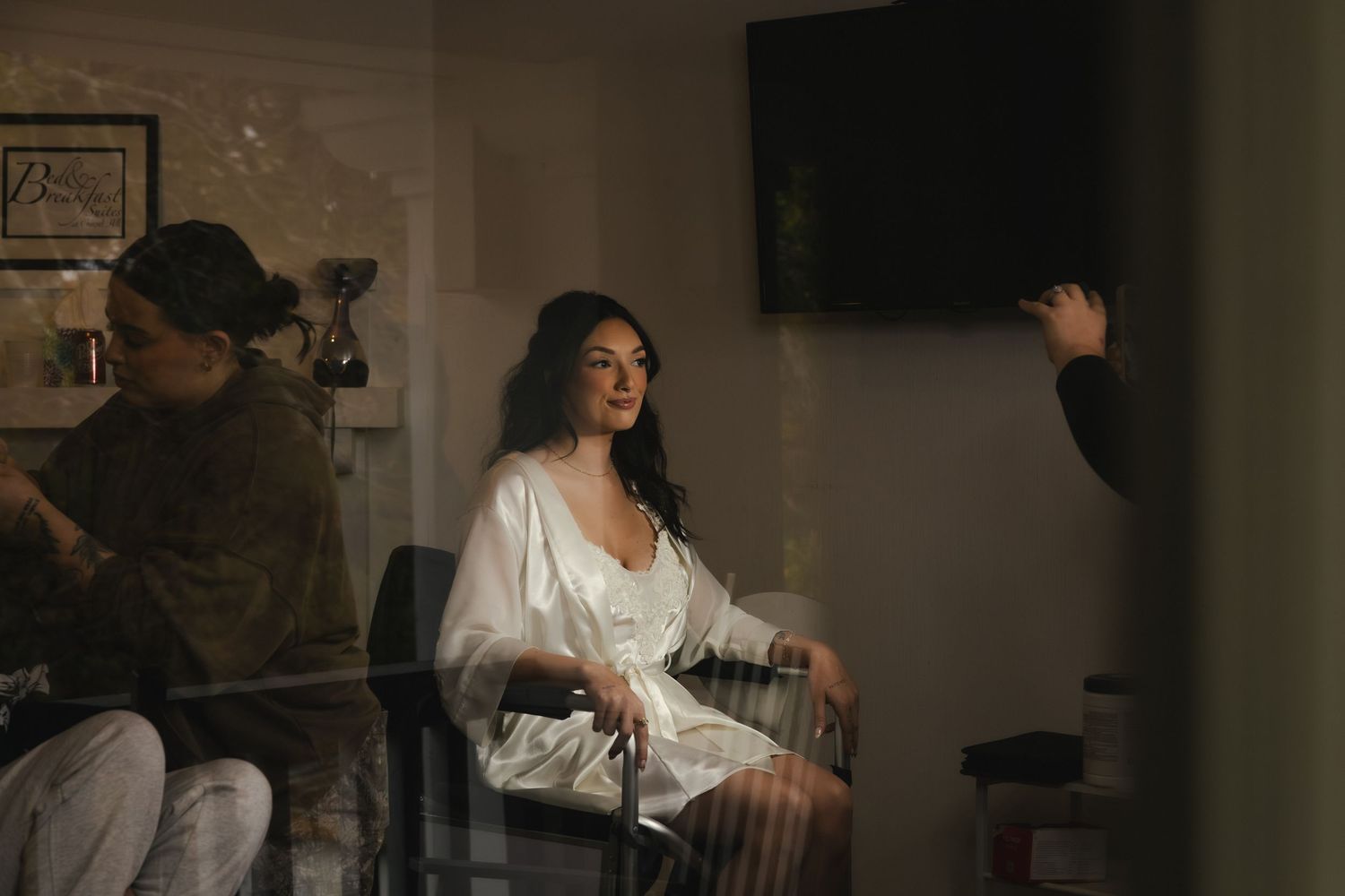 Behind-the-scenes shot of a person in a white robe being prepared for a photoshoot in a dimly lit room.