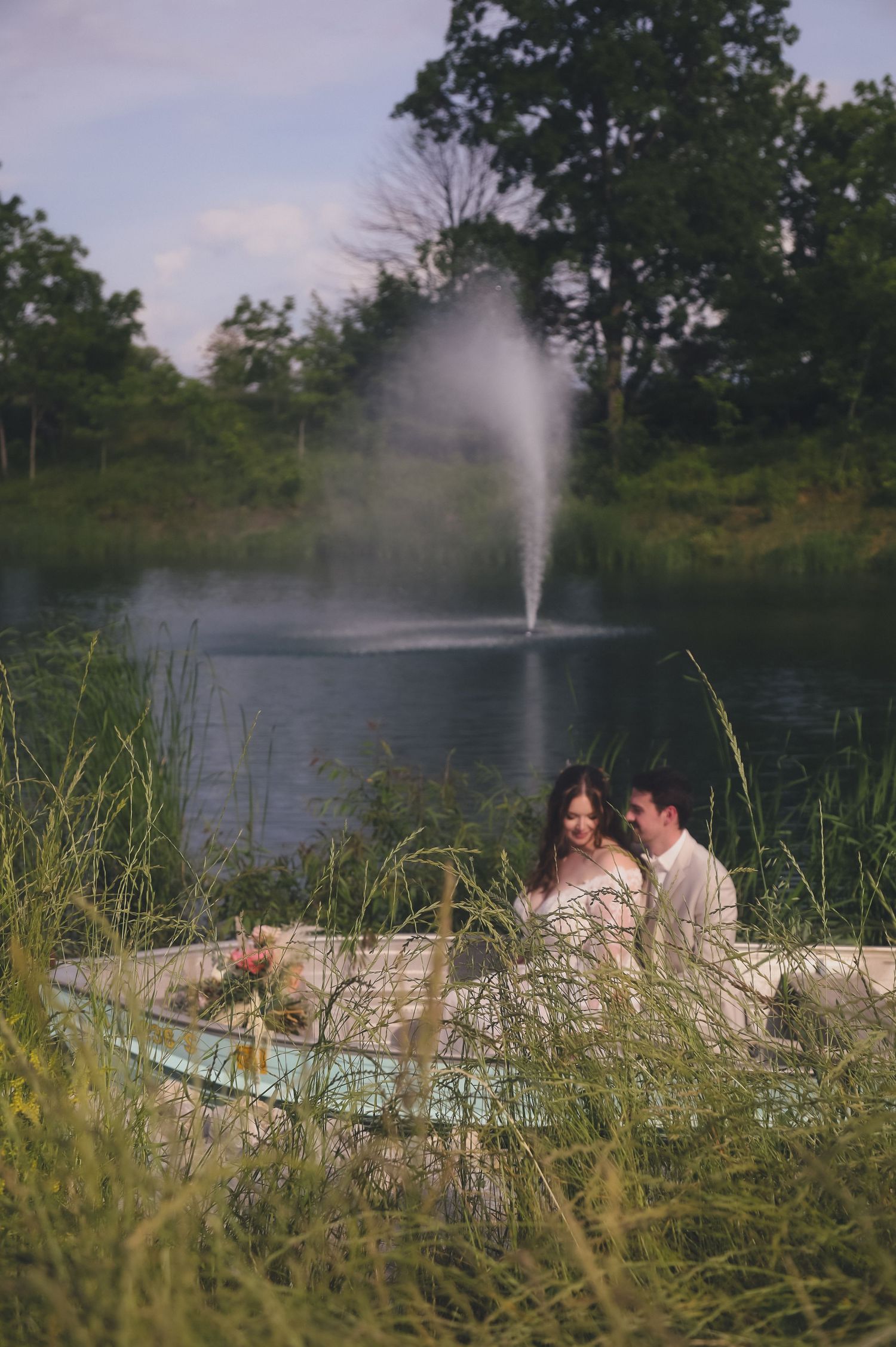 A couple relaxes on a blanket by a pond with a fountain spraying water in the background amidst tall grass at sunset.