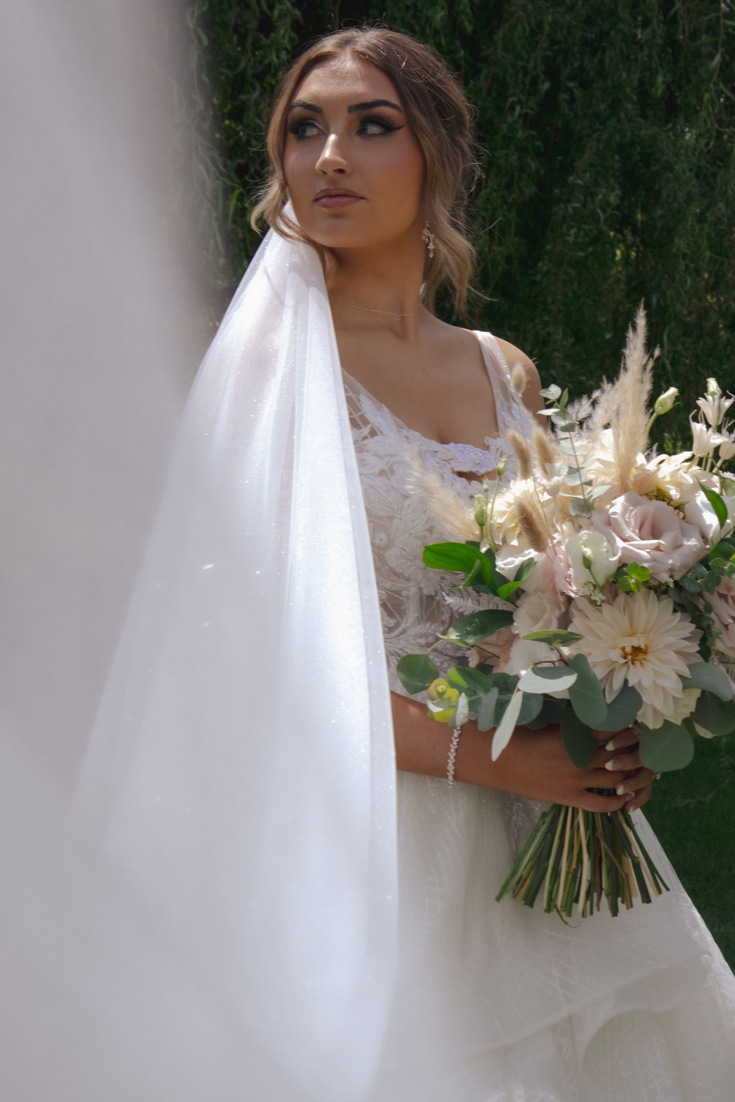Elegant bride in white gown holding neutral bouquet against green hedge backdrop.