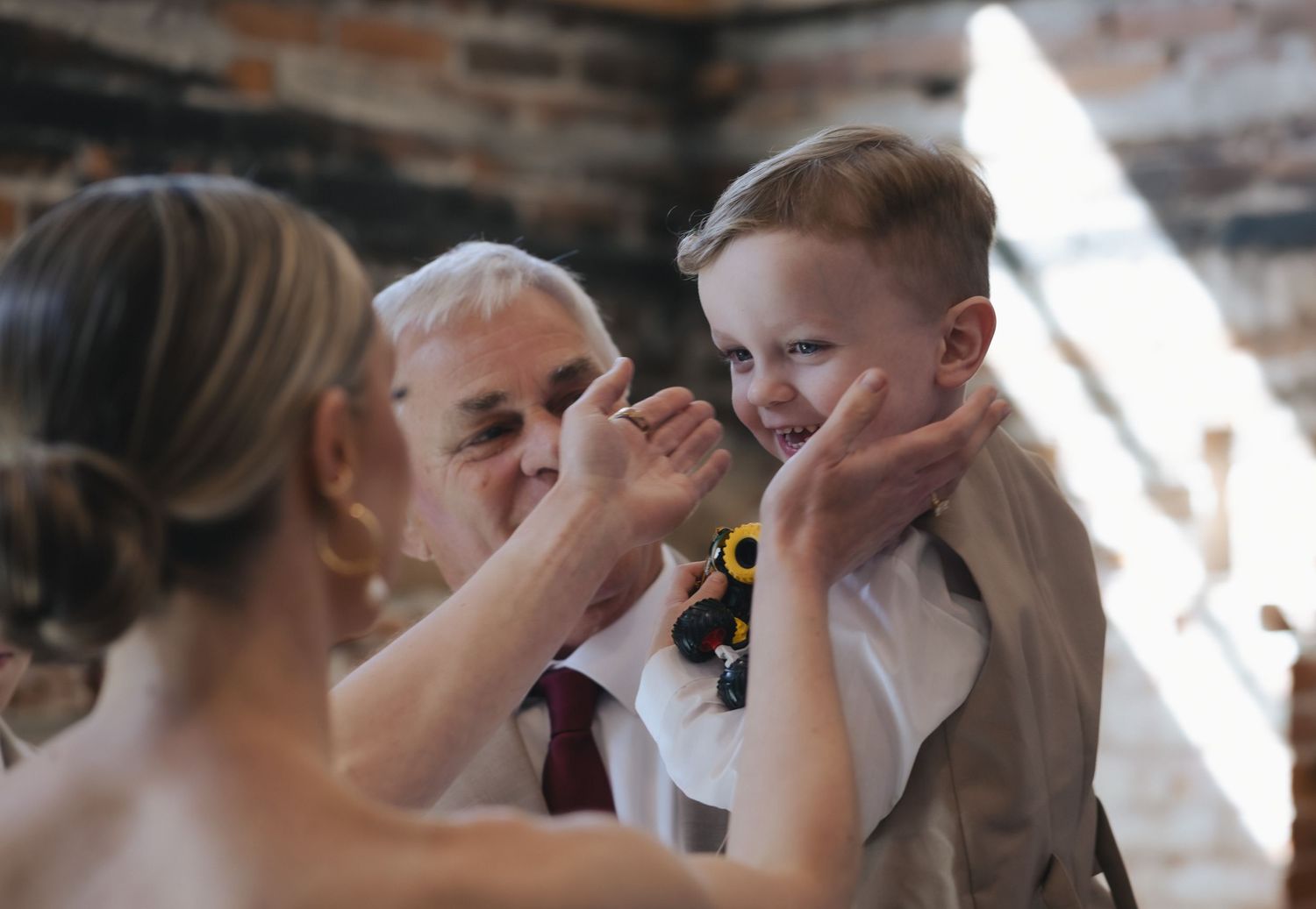 A young boy in a white shirt smiles as two adults tenderly hold his face during a wedding ceremony.