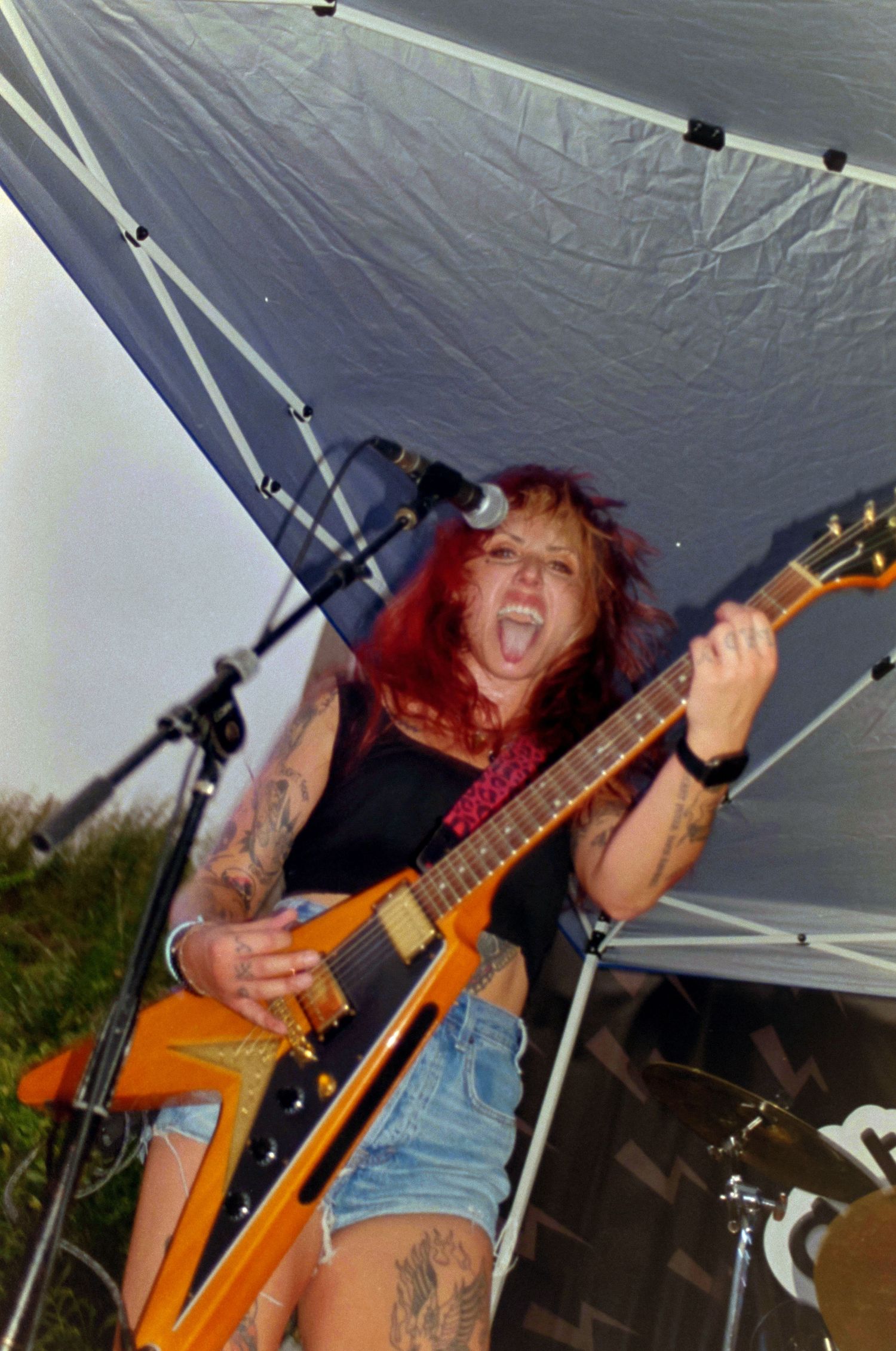 Tattooed female guitarist with red hair playing orange electric guitar on outdoor stage under tent, performing with intense energy.