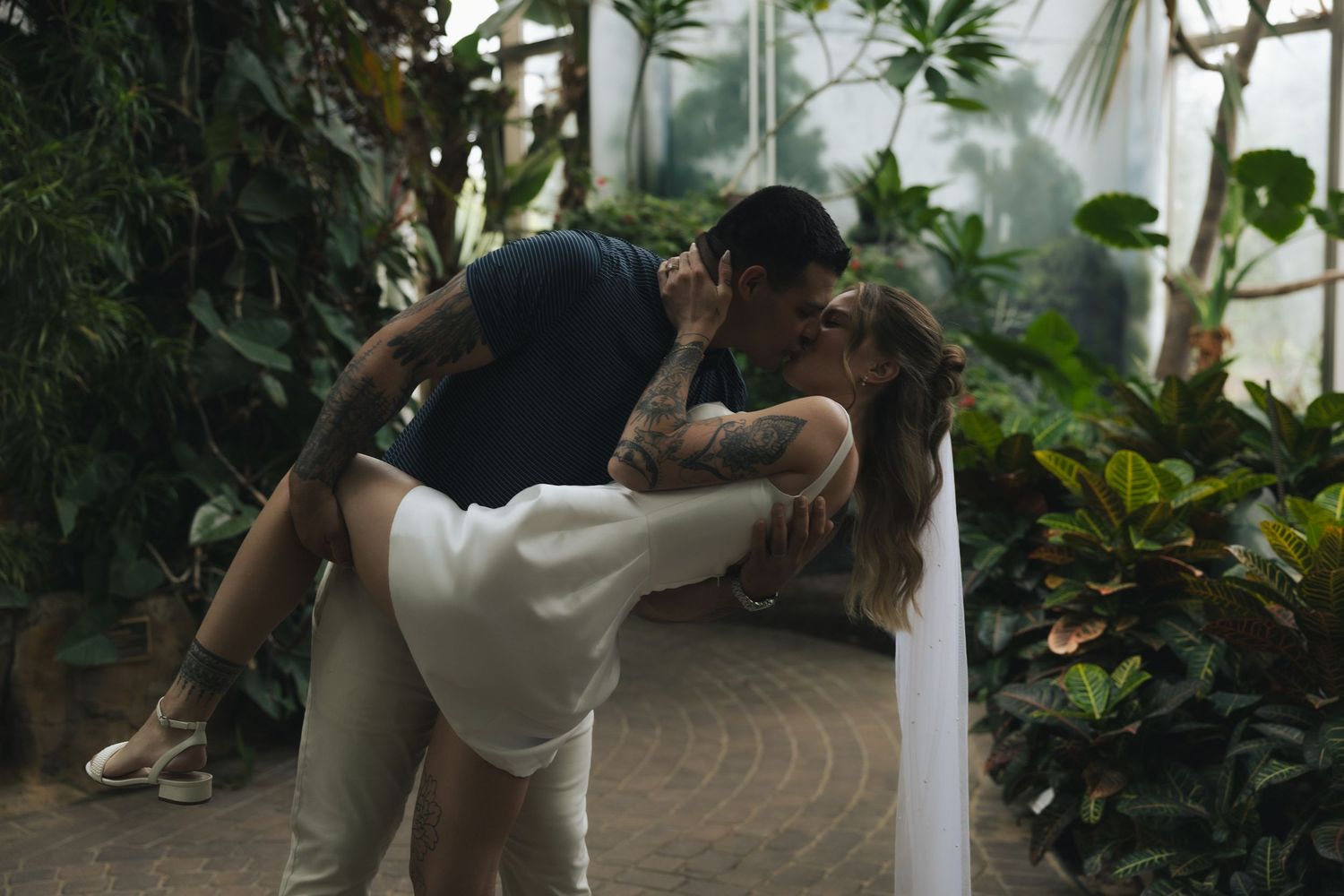 Tattooed couple sharing a passionate dip kiss surrounded by tropical plants in a greenhouse setting.