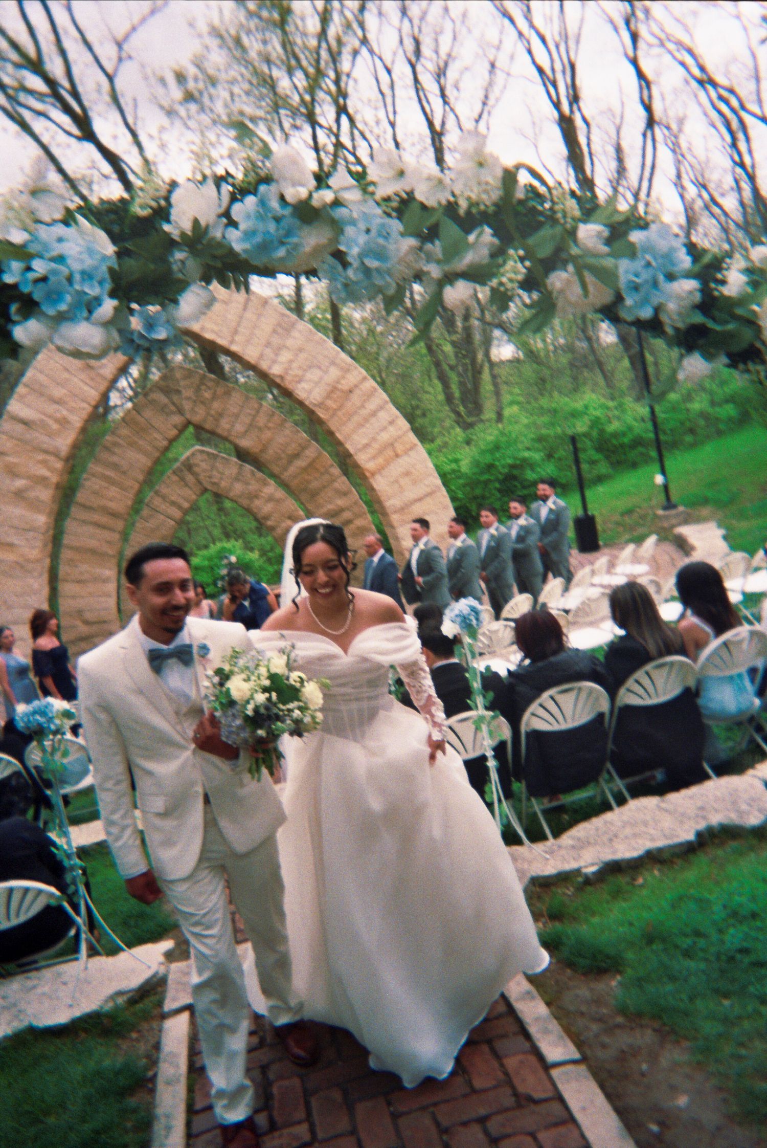 A newlywed couple walks down a brick path under an arch decorated with blue and white flowers at an outdoor ceremony.