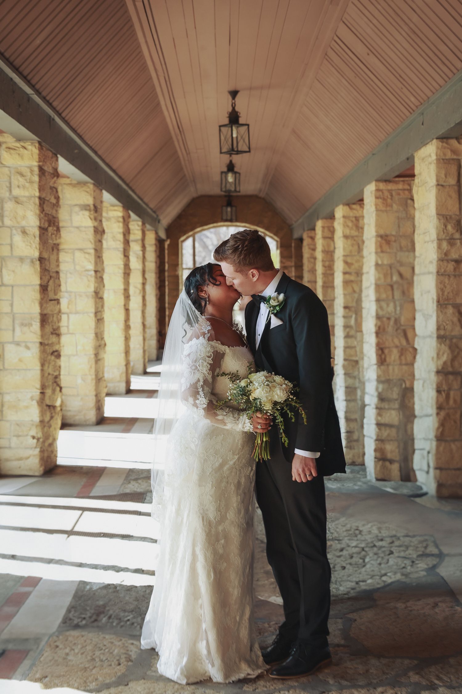 A newly married couple shares a romantic moment in a stone colonnade with arched ceilings and hanging lanterns.