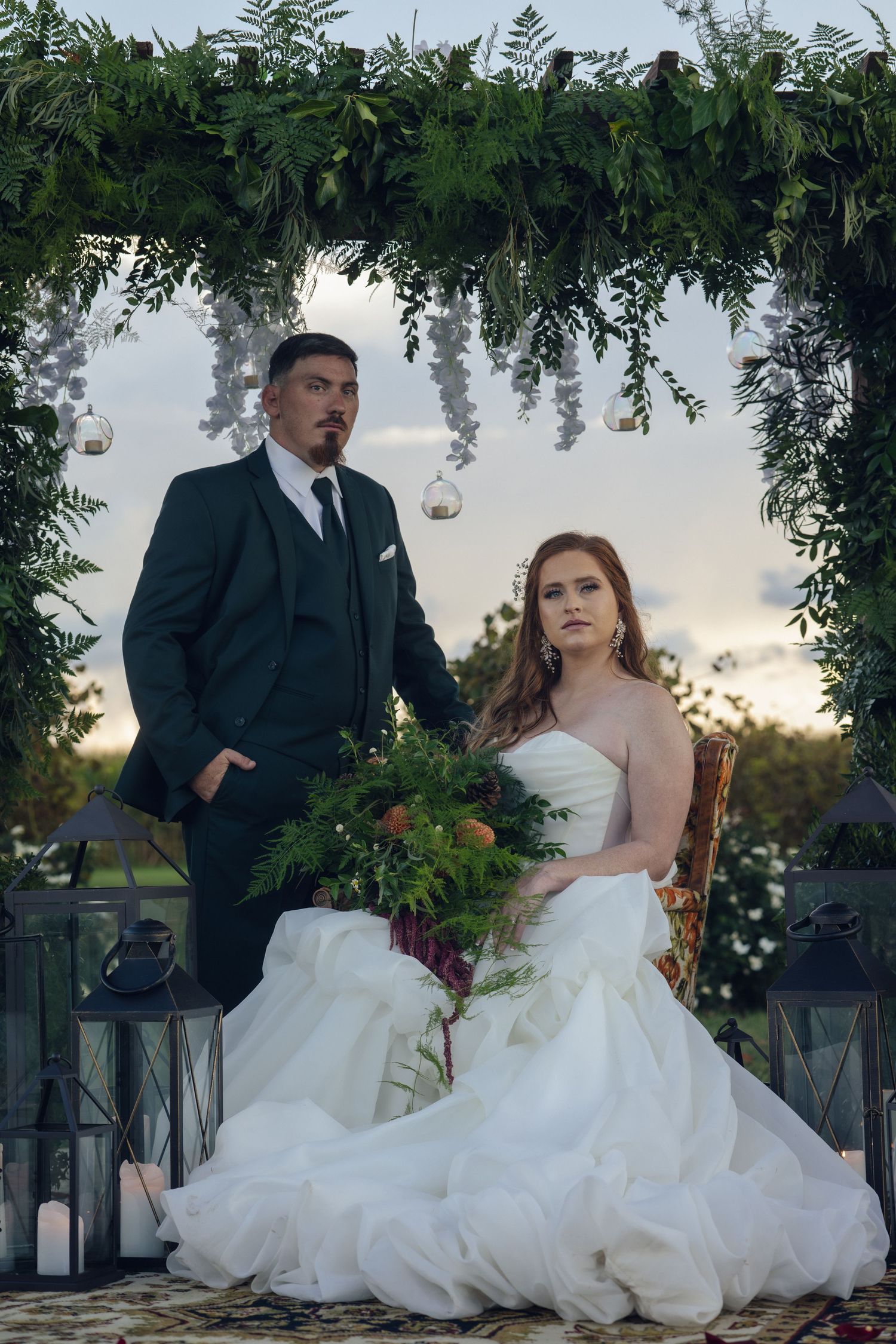 Elegant wedding couple poses under greenery arch with lanterns against sunset sky at outdoor ceremony.