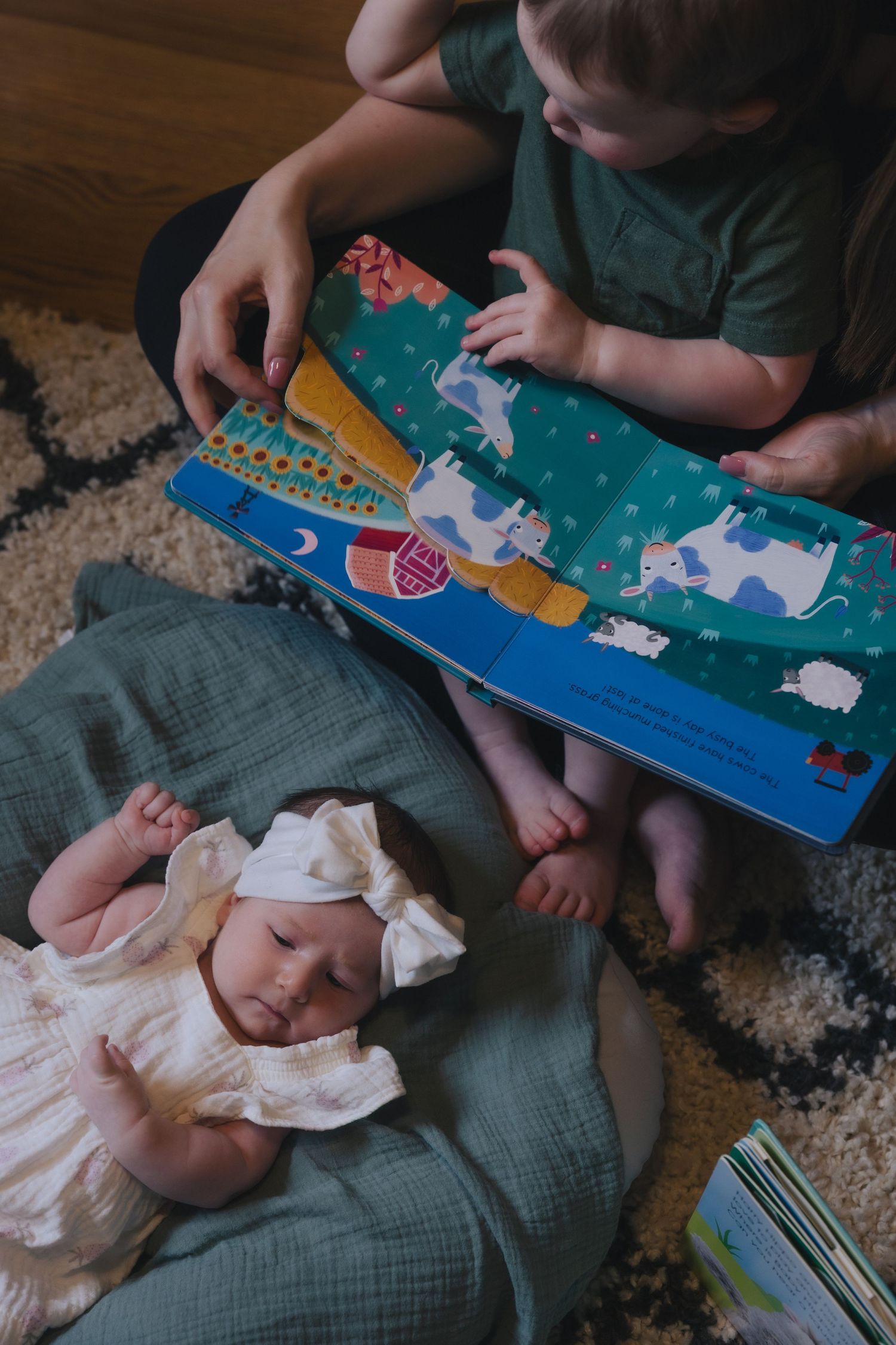 Toddler reads colorful farm animal board book beside sleeping newborn baby in white dress on soft blanket.