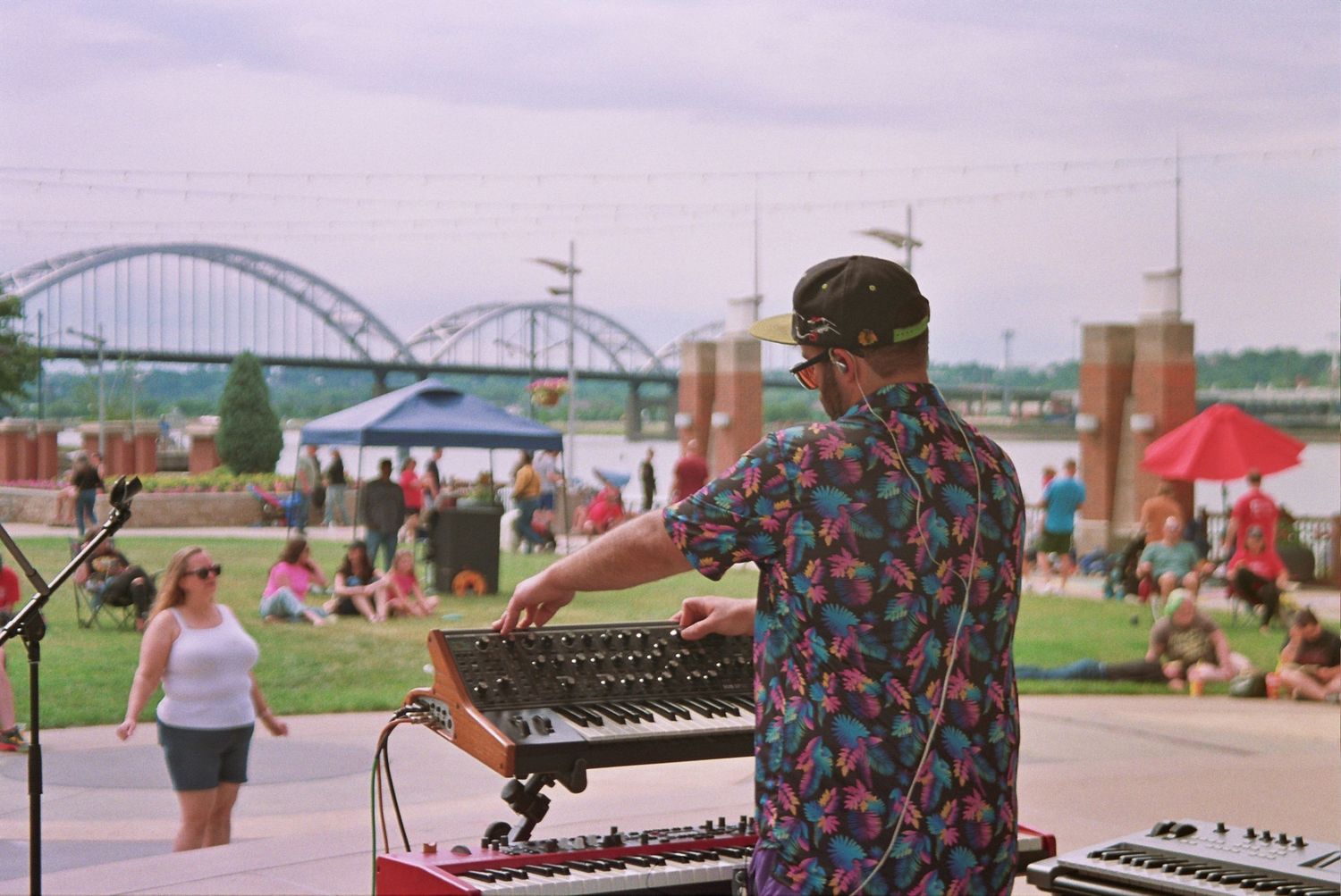 Musician playing keyboard synthesizer outdoors at a riverside festival with a bridge and crowd in the background.