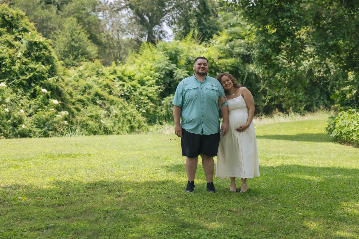 Pregnant couple standing together on green grass lawn surrounded by lush trees outdoors during summer.