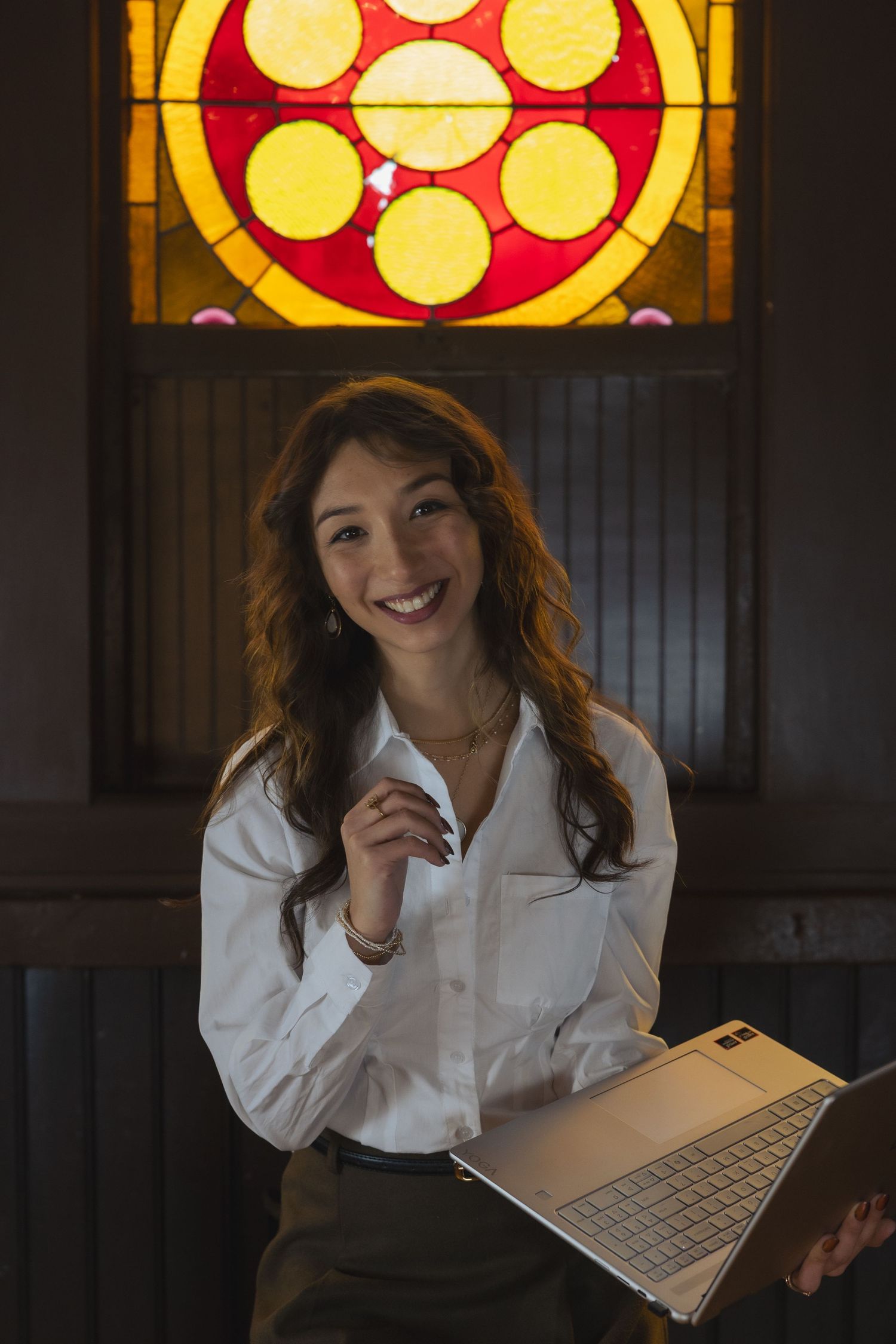 Smiling woman in white shirt holds laptop before colorful stained glass window in dark interior.