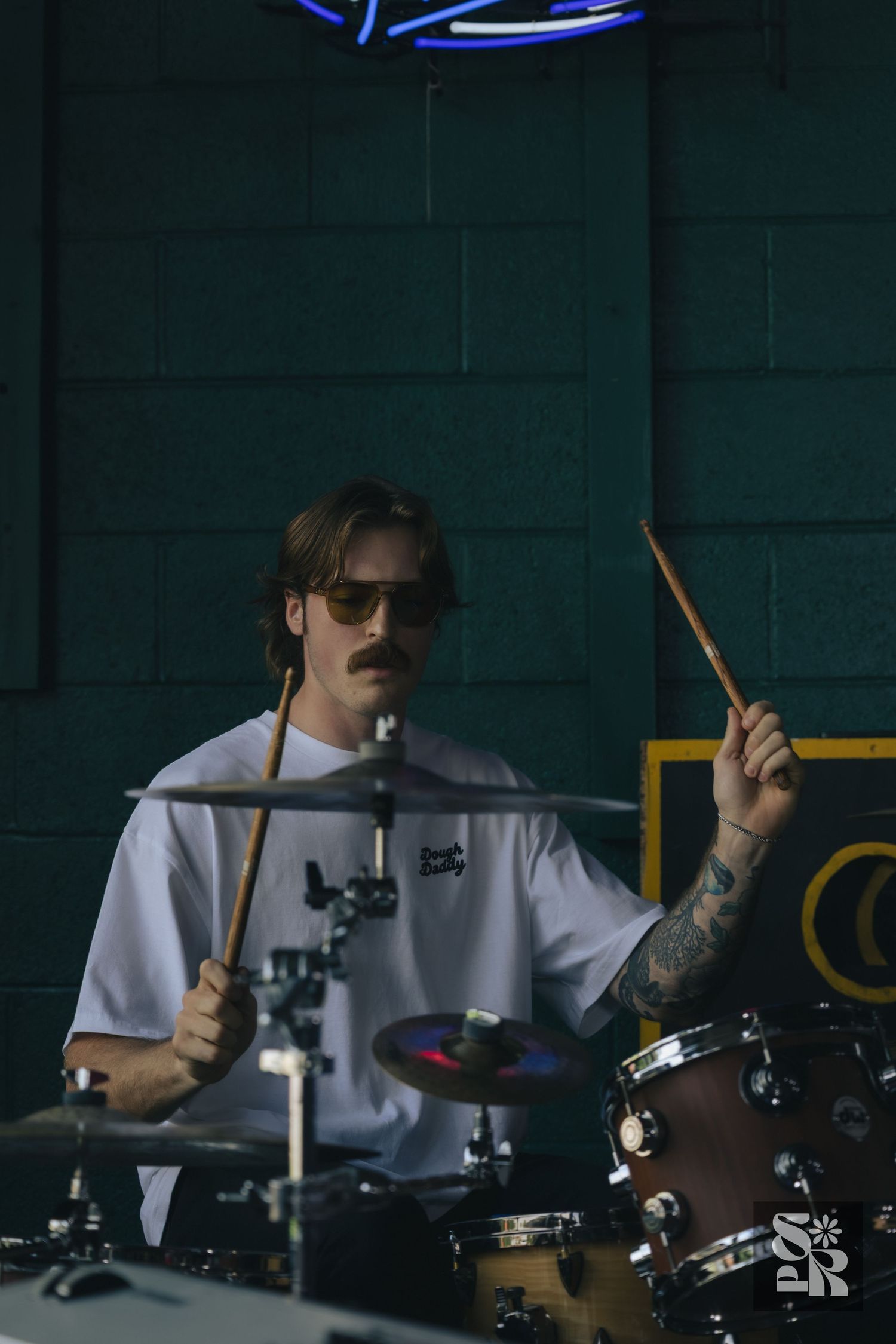 Tattooed drummer with sunglasses playing drums with sticks in a dark studio with neon lights.