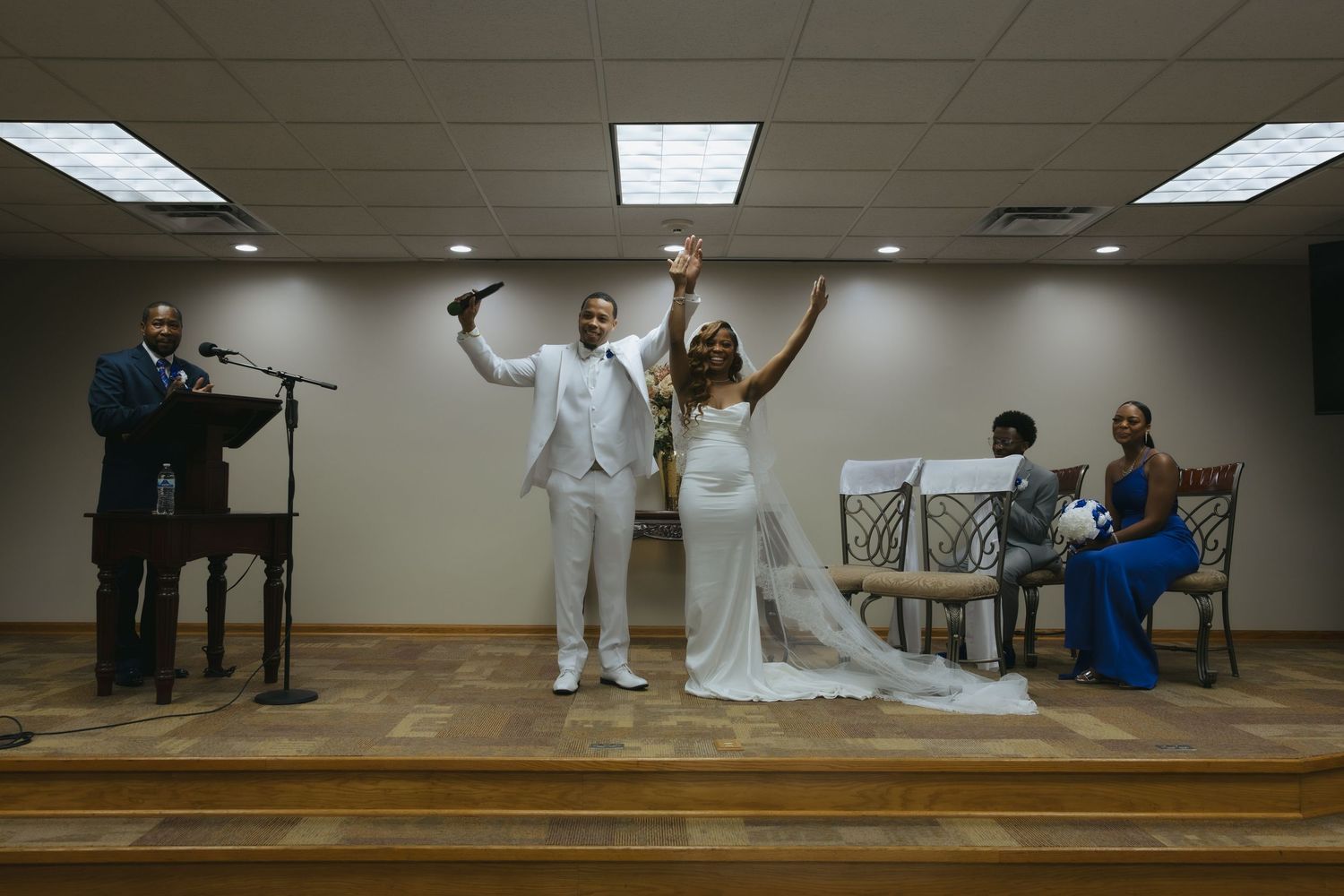 A joyful bride and groom celebrate their wedding ceremony indoors, arms raised, as an officiant speaks at a podium nearby.