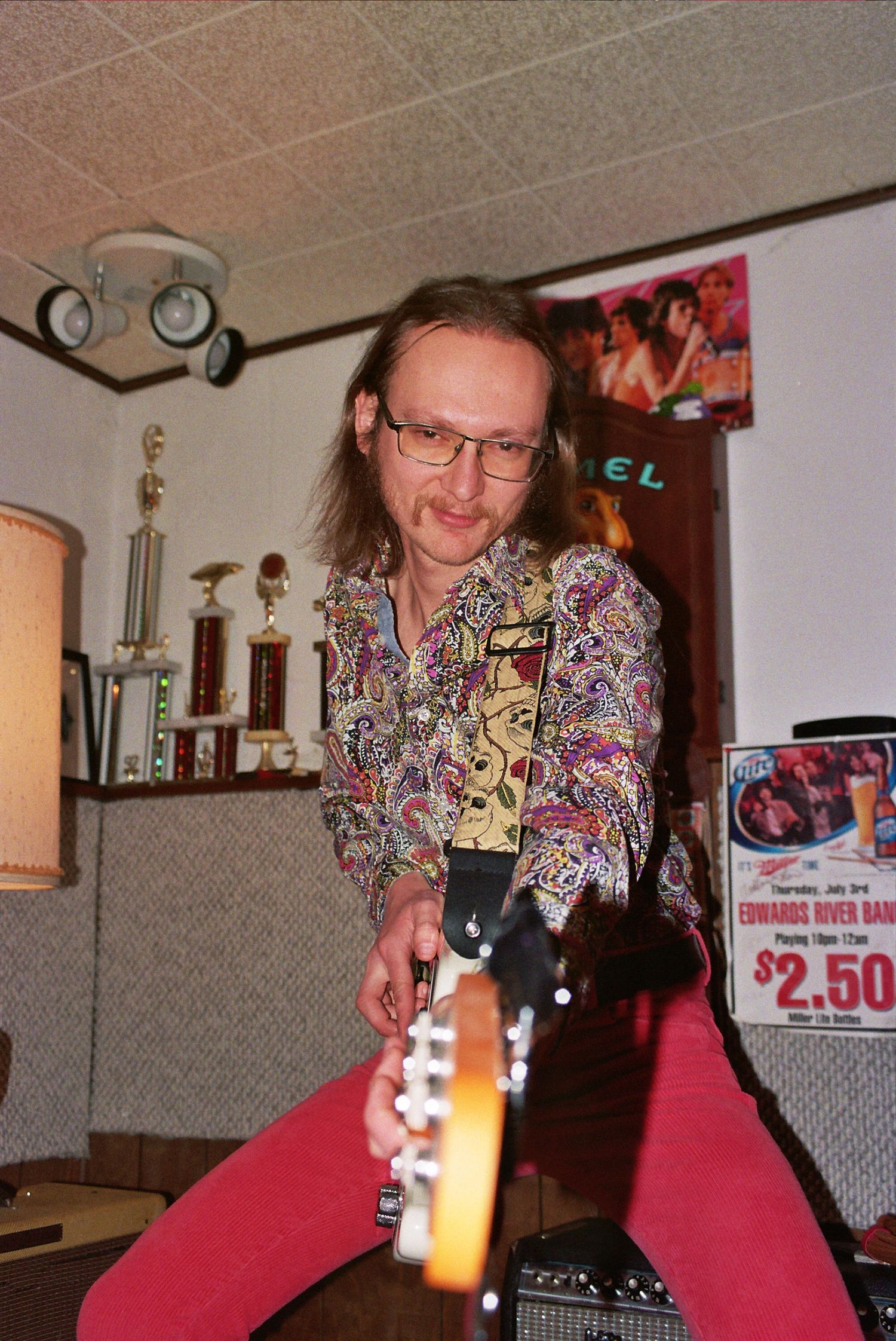 A long-haired man in a colorful paisley shirt plays an electric guitar indoors near trophies and a sign.
