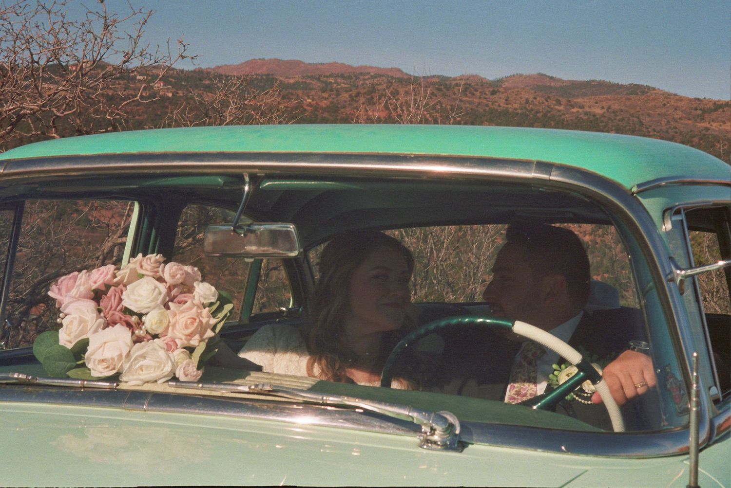 Couple in a vintage mint green car with pink roses bouquet, desert mountains backdrop, wedding day.