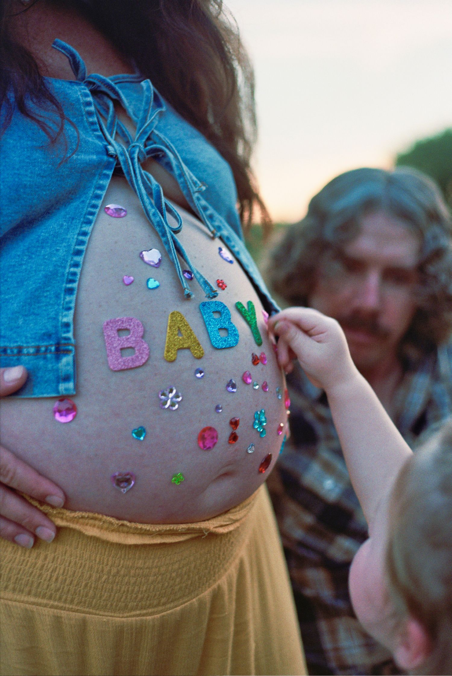 A pregnant belly decorated with colorful letter stickers spelling 'BABY' and sparkly embellishments under denim clothing.