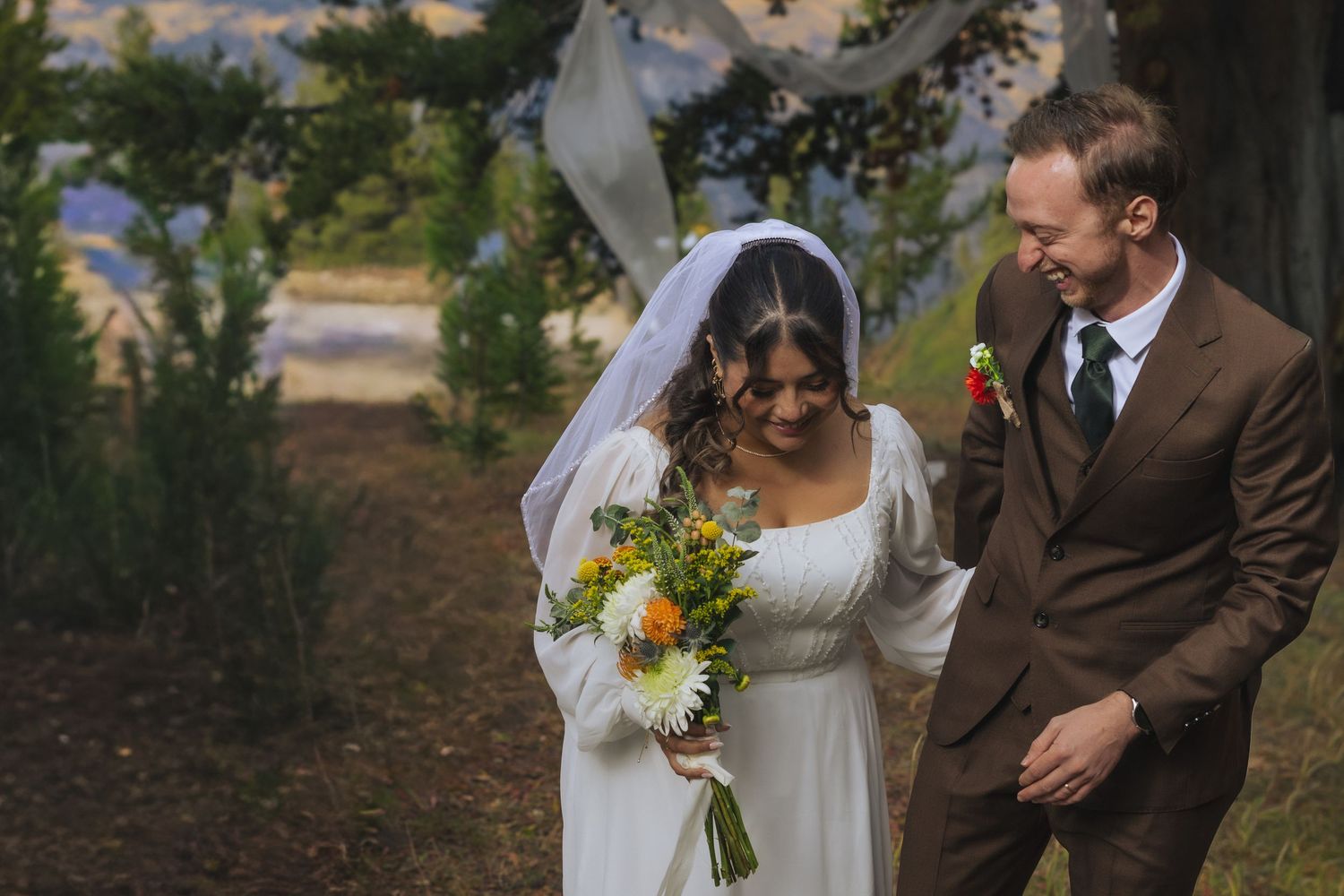 Couple shares joyful moments during outdoor wedding ceremony with sunflowers and rustic woodland backdrop.