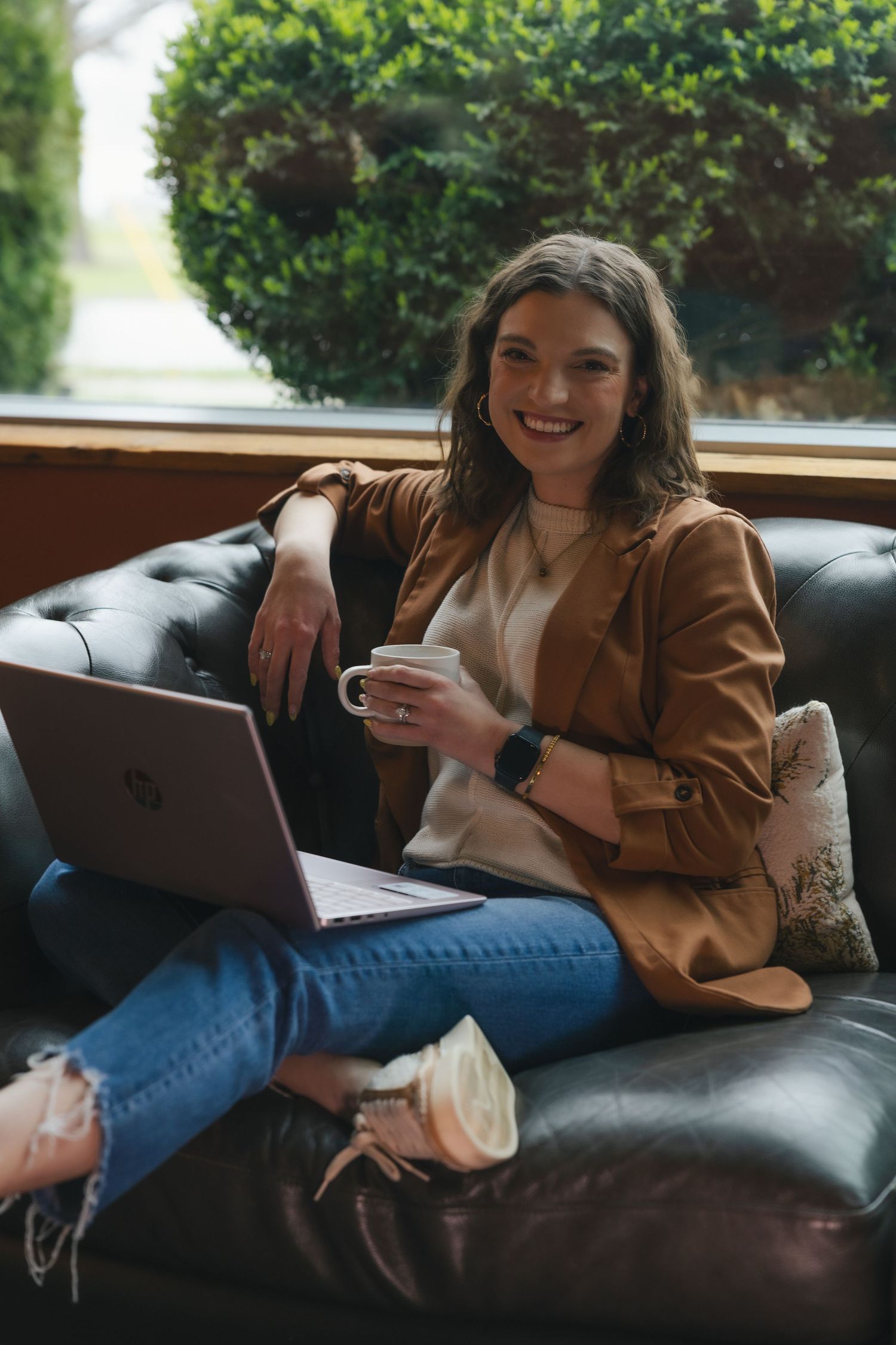 Smiling woman in brown blazer sits on leather sofa with HP laptop and coffee cup near window.