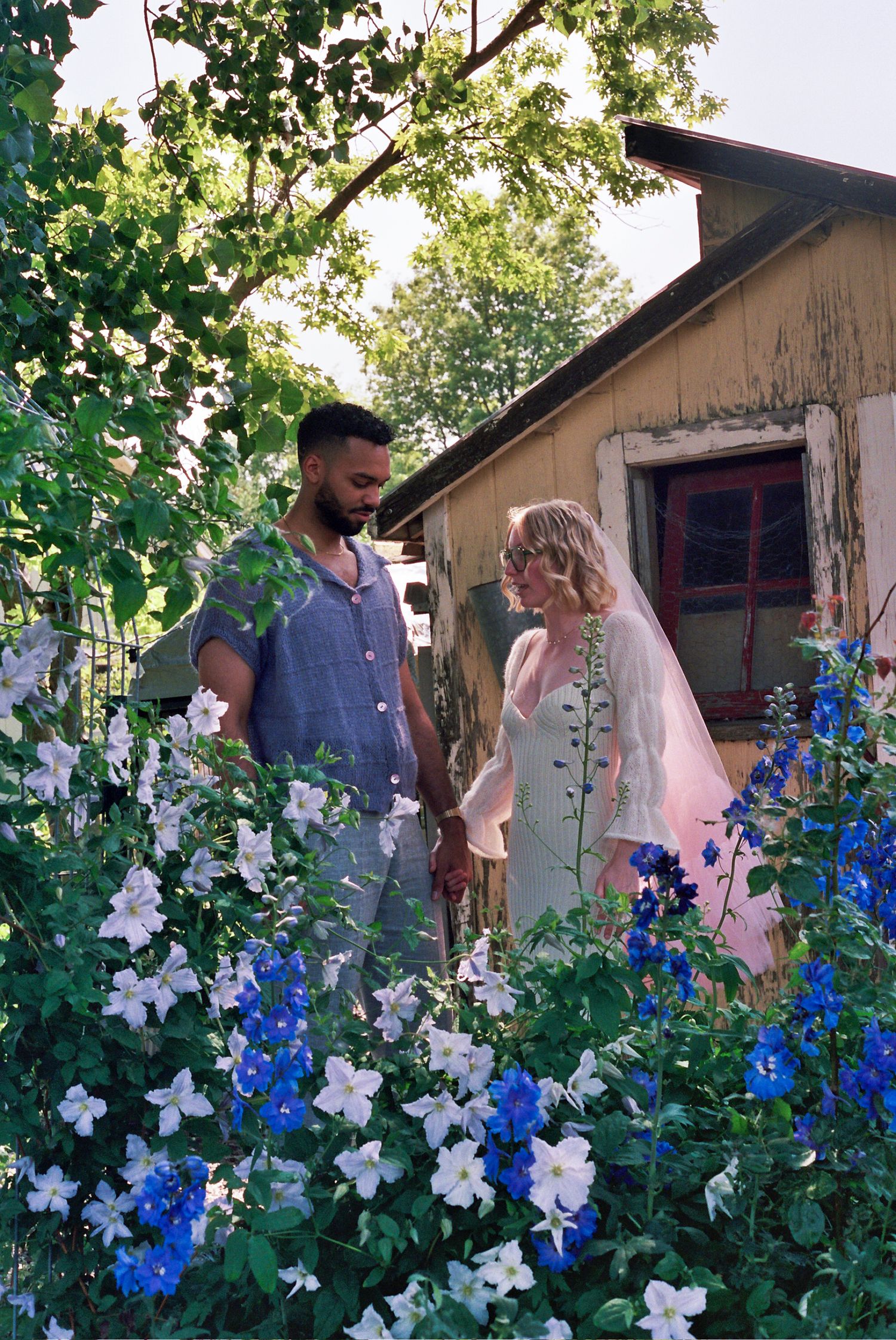 A couple stands in front of a rustic cabin surrounded by vibrant blue and white delphinium flowers in a garden setting.