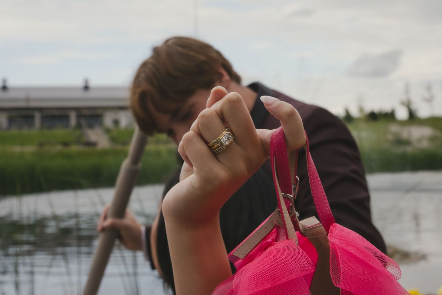 A close-up shot of a hand wearing a ring and holding a bright pink purse handle near a lake.