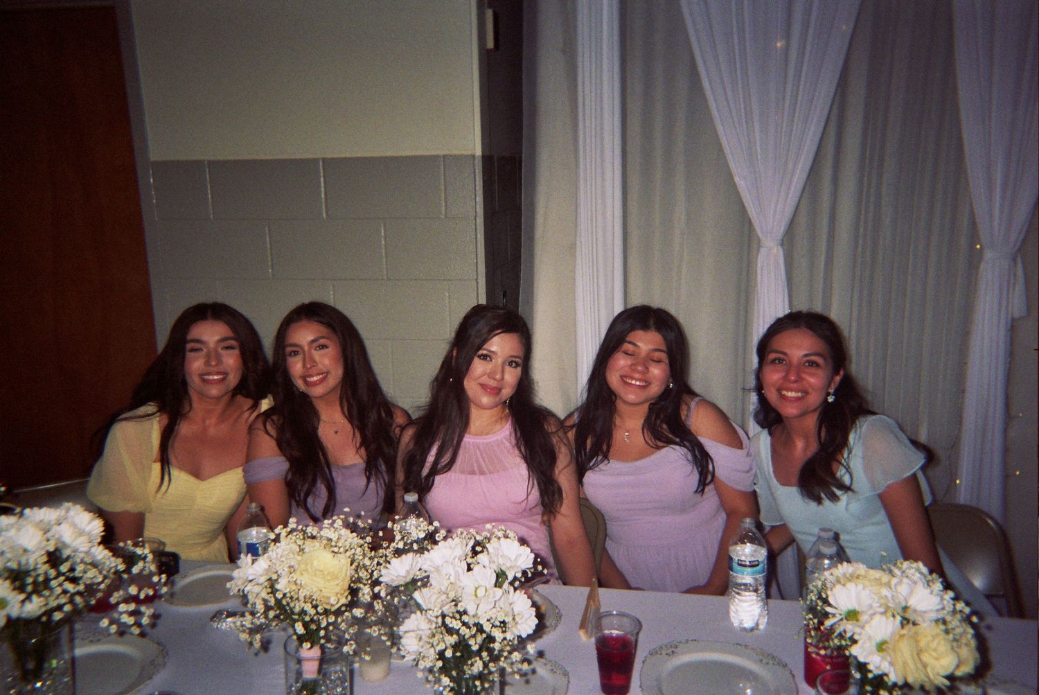 A group gathers around white floral bouquets at a wedding reception celebration.