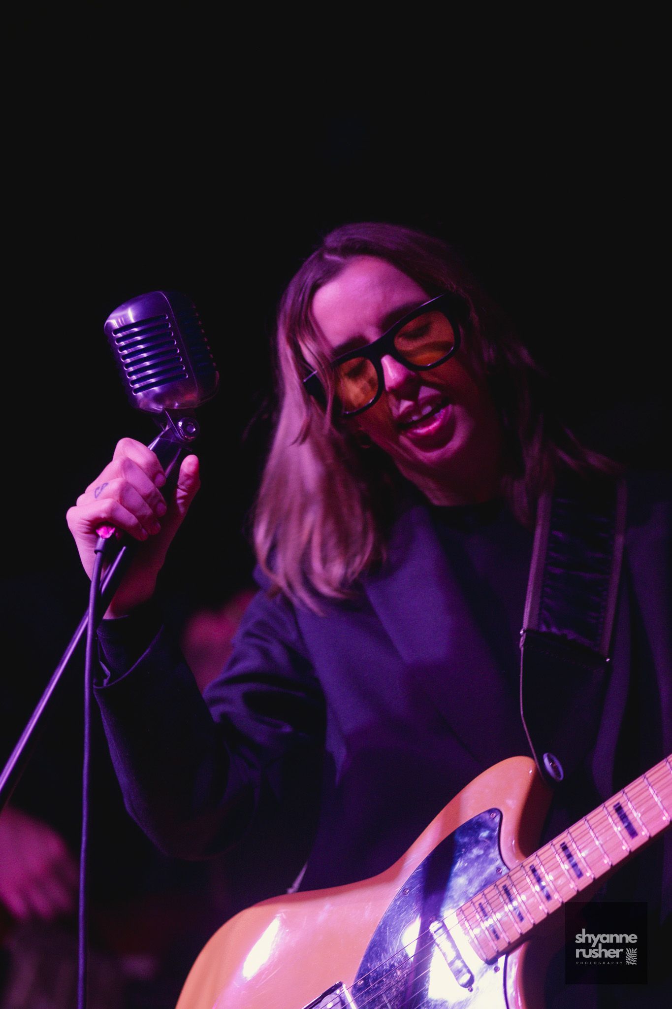 A performer in purple lighting sings passionately into a vintage microphone while holding an acoustic guitar on stage.