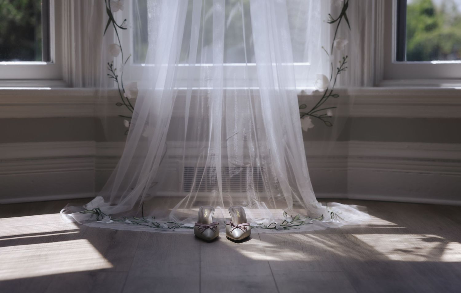 Delicate white wedding veil draped over bridal shoes by a sunlit window with sheer curtains.