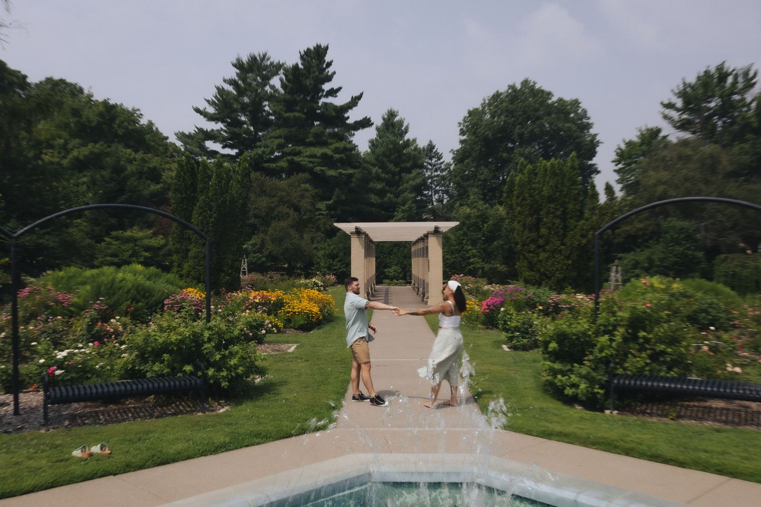 Couples enjoying a beautiful garden park with a decorative fountain, lush greenery, colorful roses, and a pergola in the background.