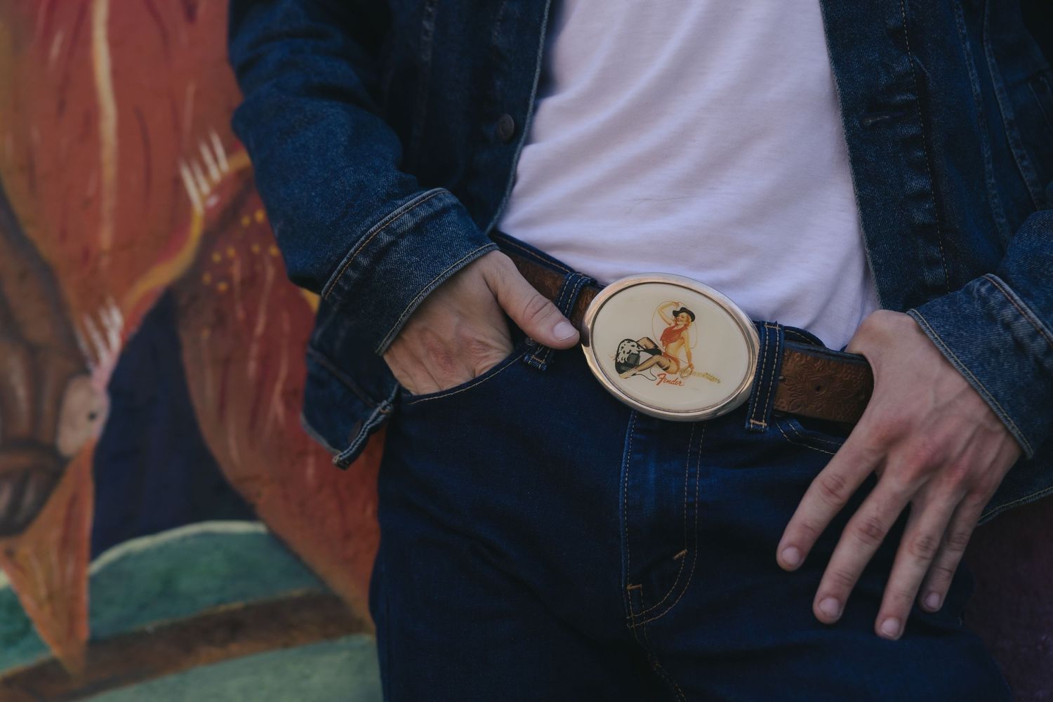 Man wearing denim jacket and jeans with a decorative oval belt buckle featuring a bird design.