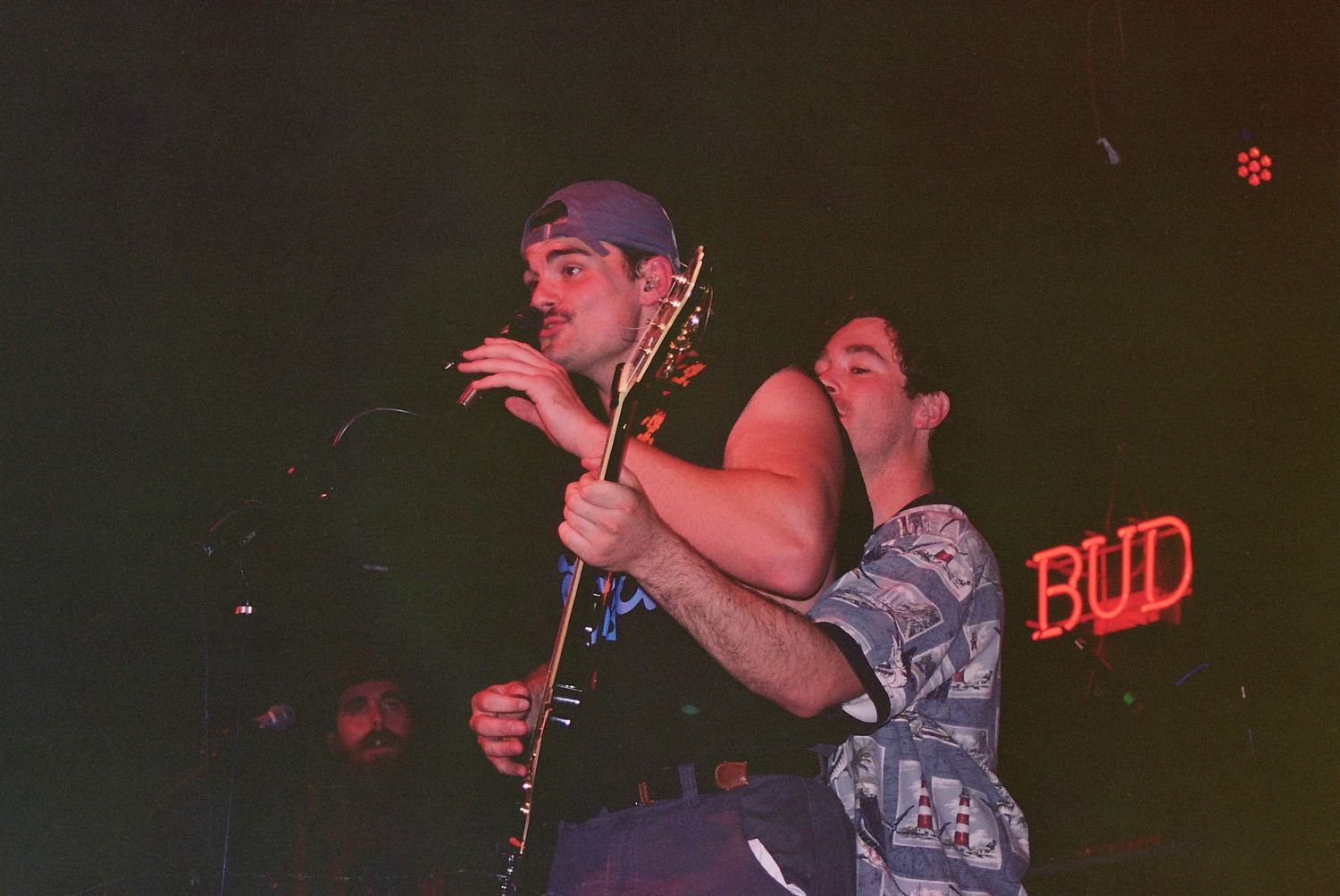 Two musicians performing on stage at a bar with a Budweiser sign glowing in the background.