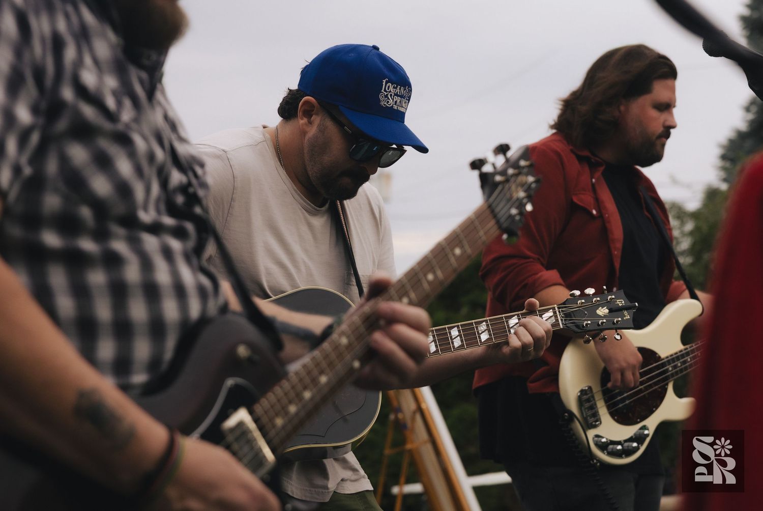 Musicians playing electric guitars outdoors at a casual music event, with one wearing a blue cap.