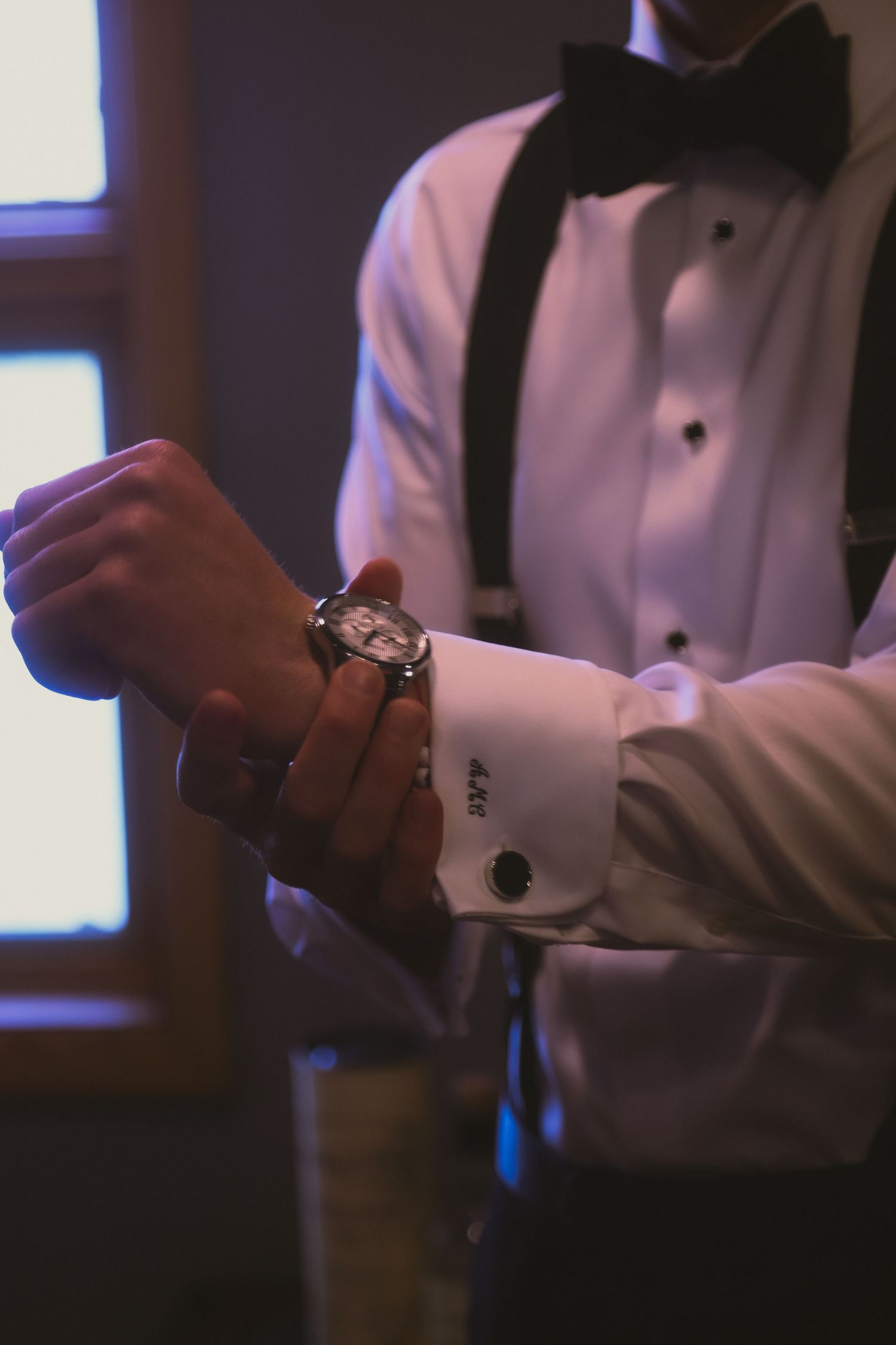 Close-up shot of formal attire showing white dress shirt sleeve with cufflink, black bow tie and wristwatch in dim lighting.