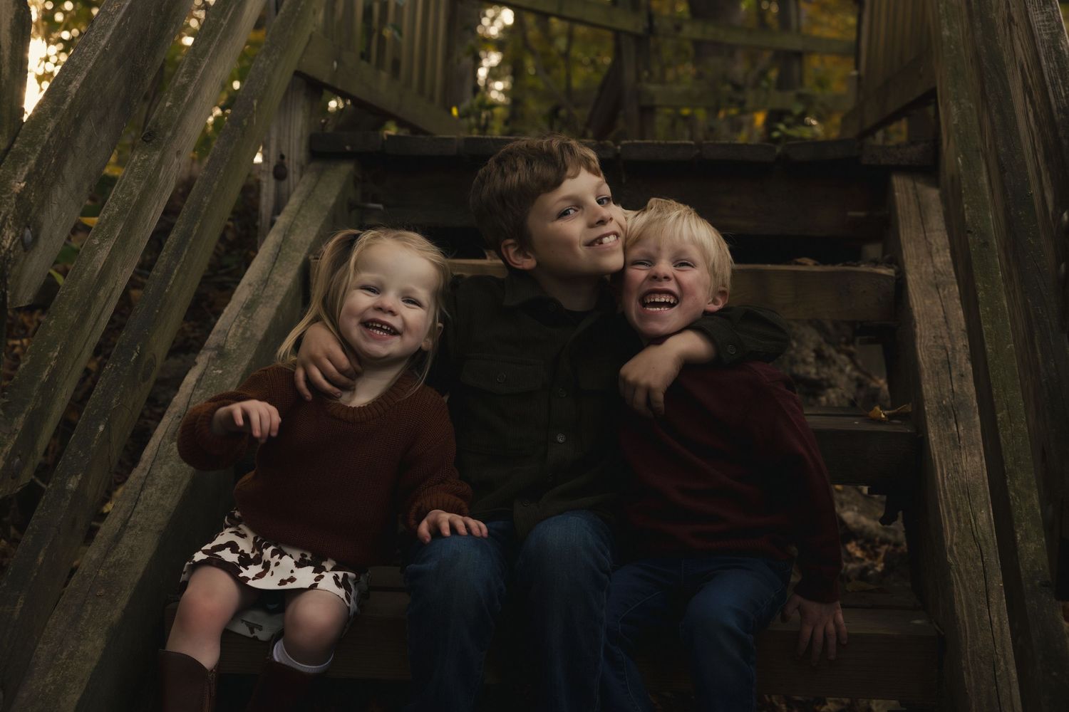Three happy children sitting on rustic wooden steps outdoors in autumn, wearing coordinating burgundy and dark outfits.