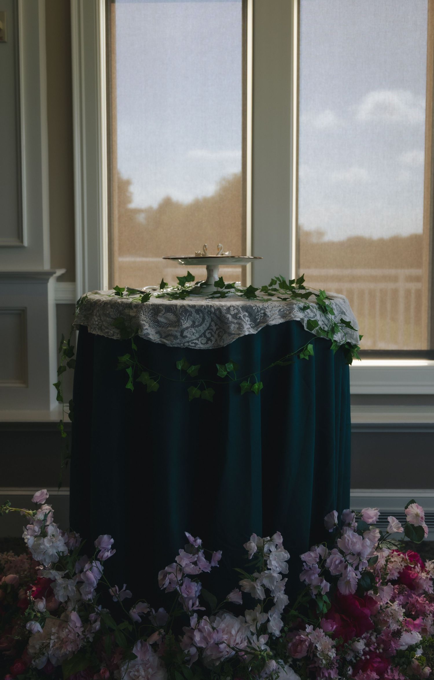 Elegant wedding cake table with dark teal linen, lace overlay, ivy garland, and pink floral arrangements near a sunlit window.
