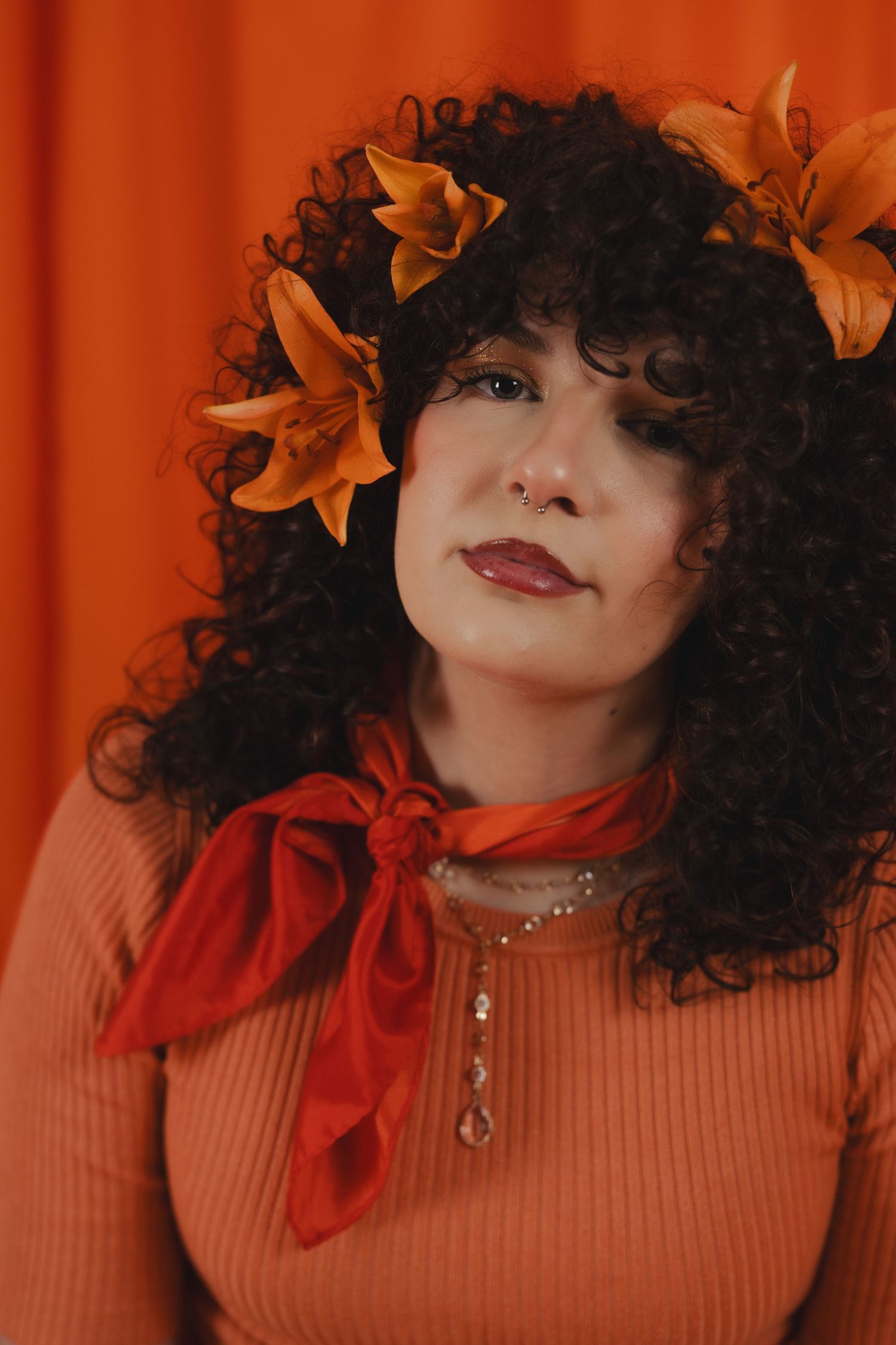 Woman with curly hair adorned with orange lilies wearing a coral sweater and red bow scarf against orange backdrop.