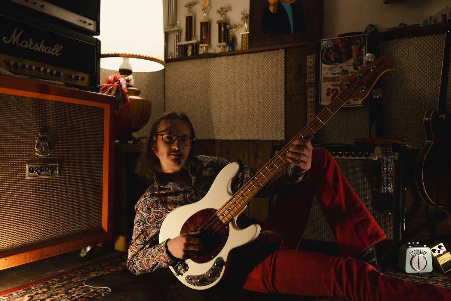 Young musician with glasses playing a white bass guitar beside Marshall amp in a cozy, dimly lit music room.
