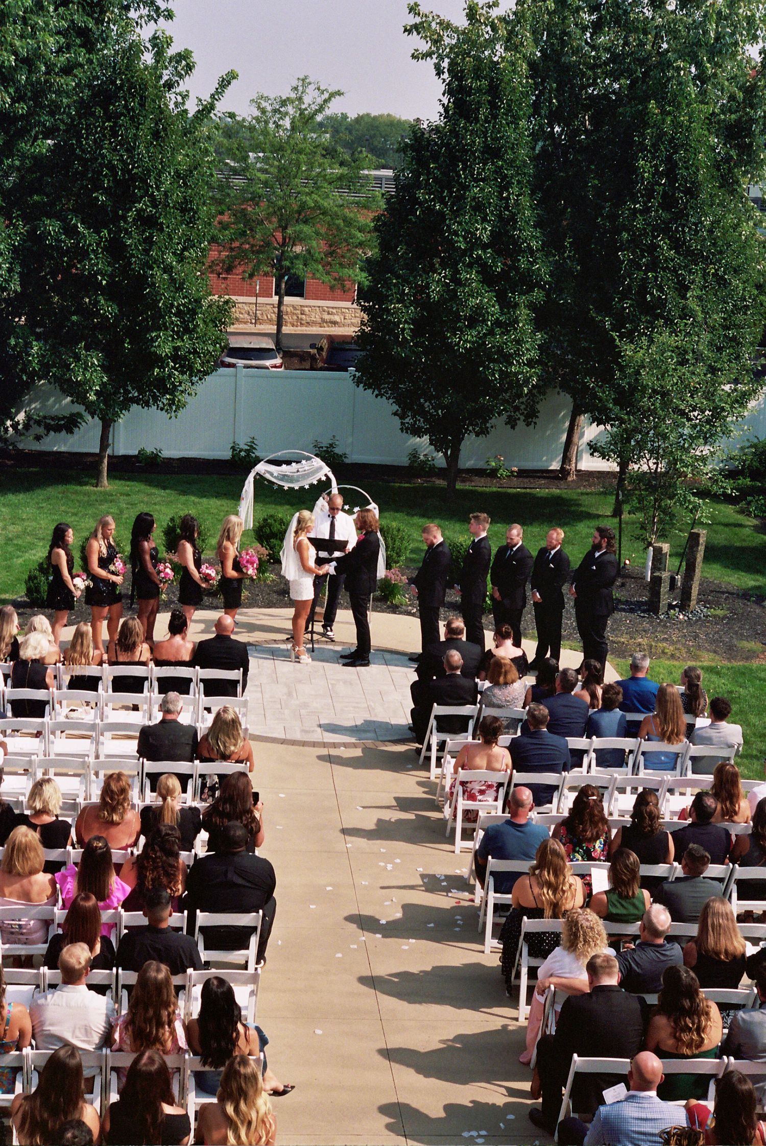 Outdoor wedding ceremony setup with white chairs arranged in rows on a patio surrounded by green trees.