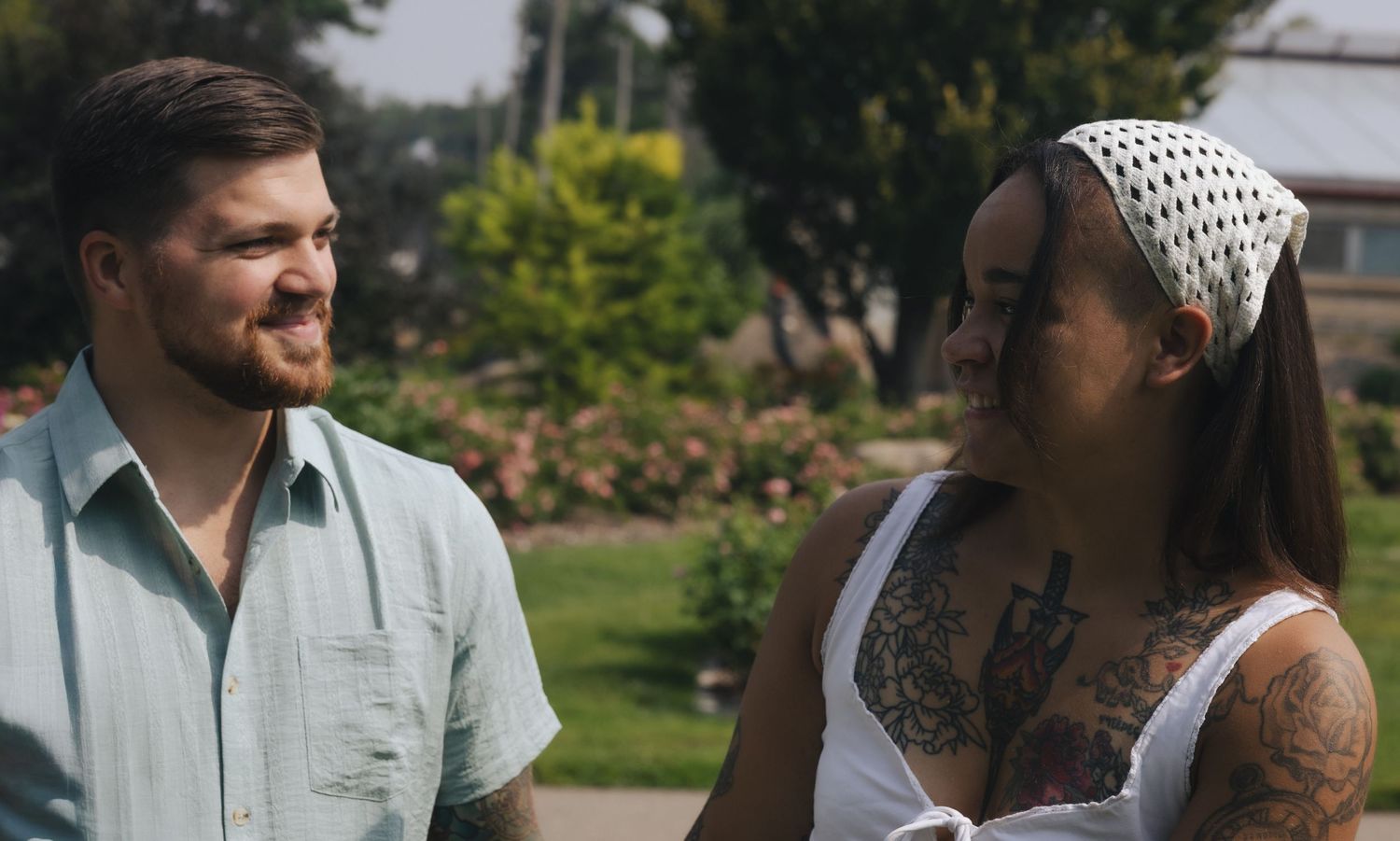 A tattooed woman in a white top and headband smiles at a man in a green shirt outdoors.