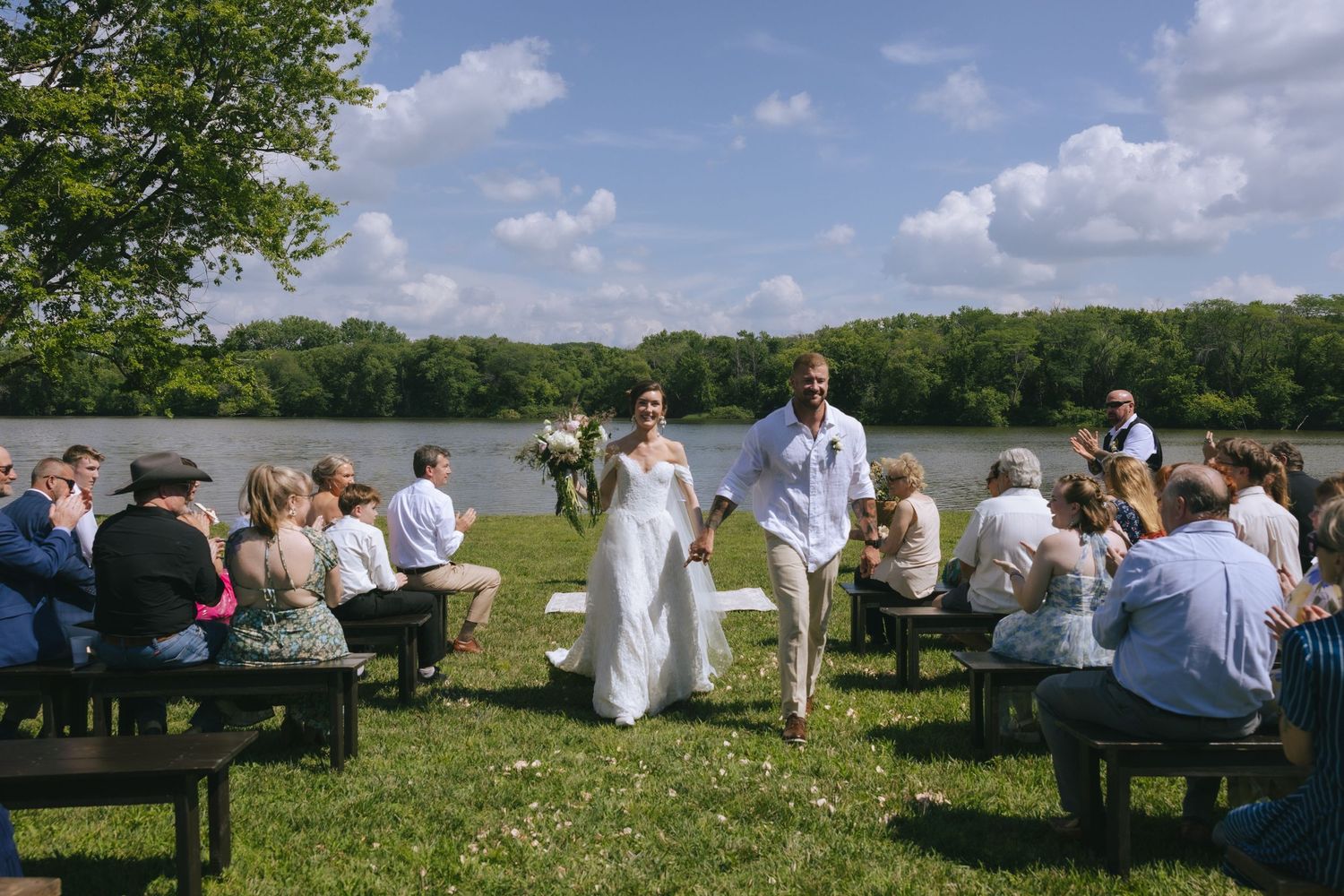 Bride and groom walk down outdoor aisle by lakeside, guests seated on wooden benches, lush trees and blue sky backdrop.