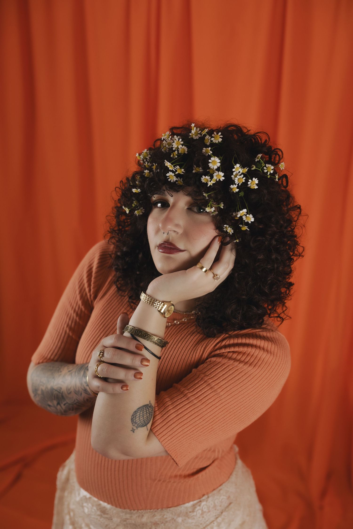 Curly-haired woman with flowers in hair, wearing orange ribbed sweater, gold watch, and tattoos, posing against red backdrop.