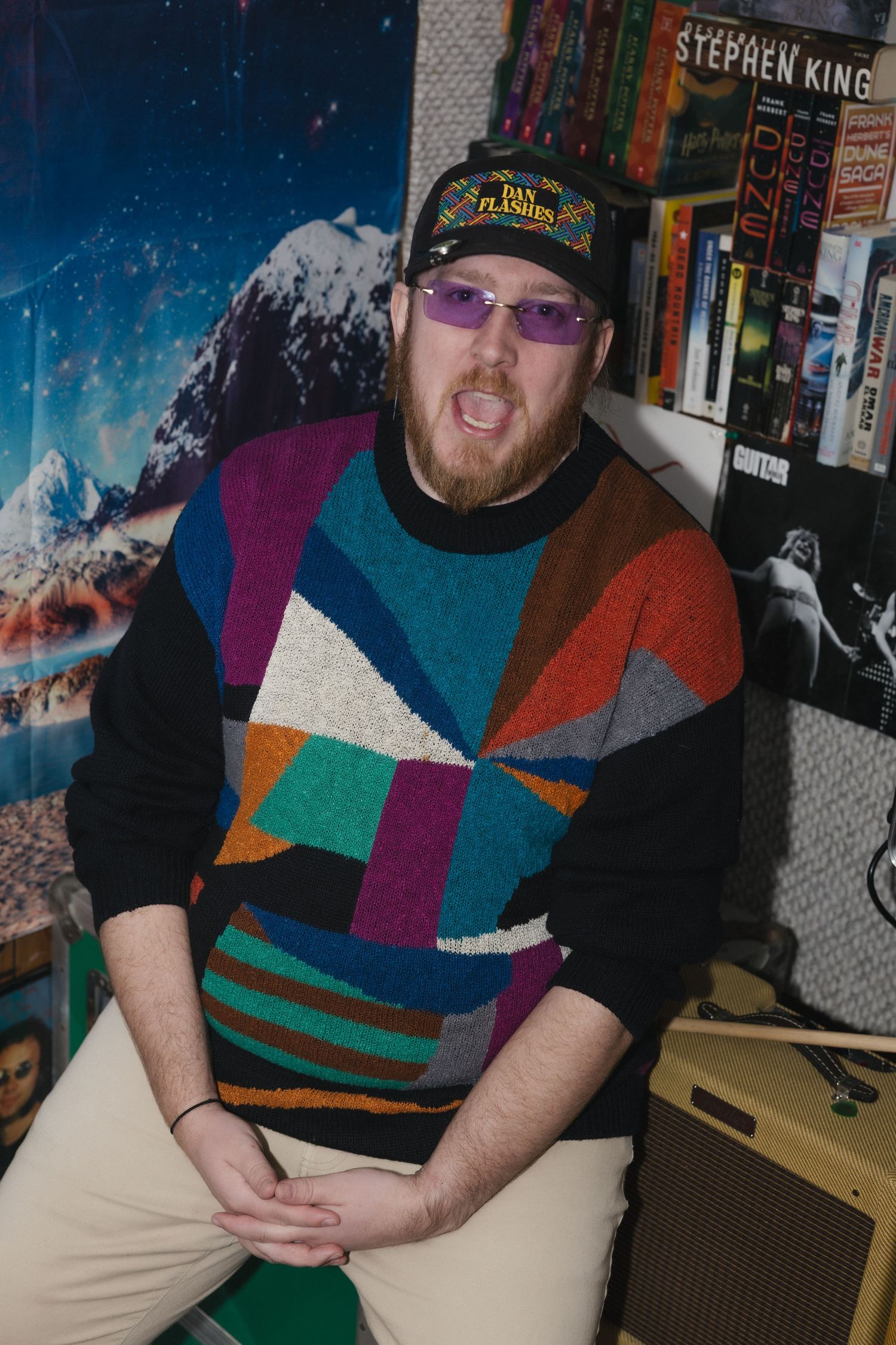 Man in colorful geometric sweater and cap sits in a room decorated with Stephen King books and mountain poster.