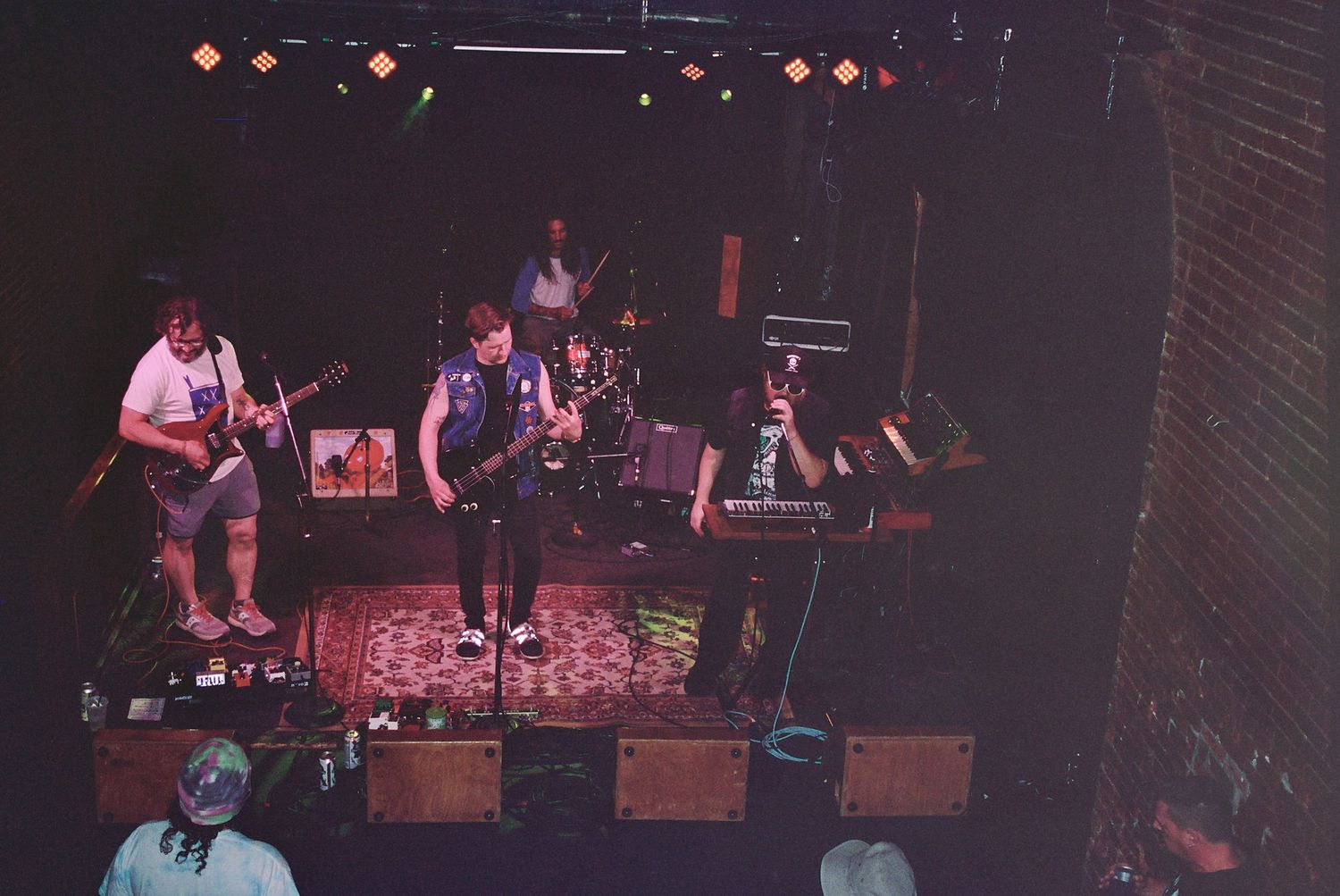 Band performing on a dimly lit stage with guitars and drums, viewed from above in a small music venue.