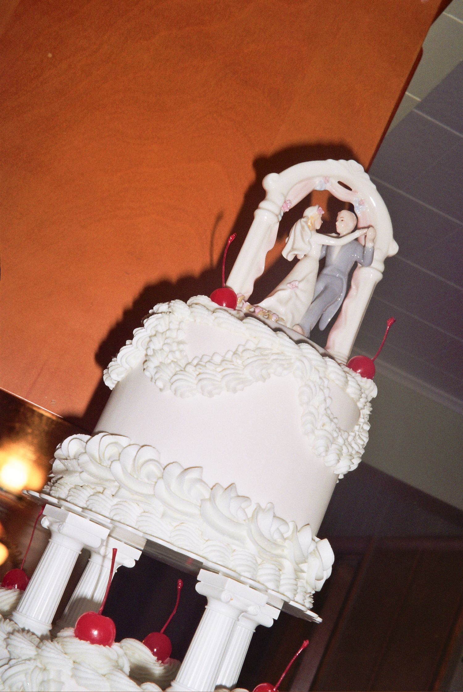 Elegant white multi-tiered wedding cake with bride and groom topper, decorative frosting, pillars, and red cherry accents.