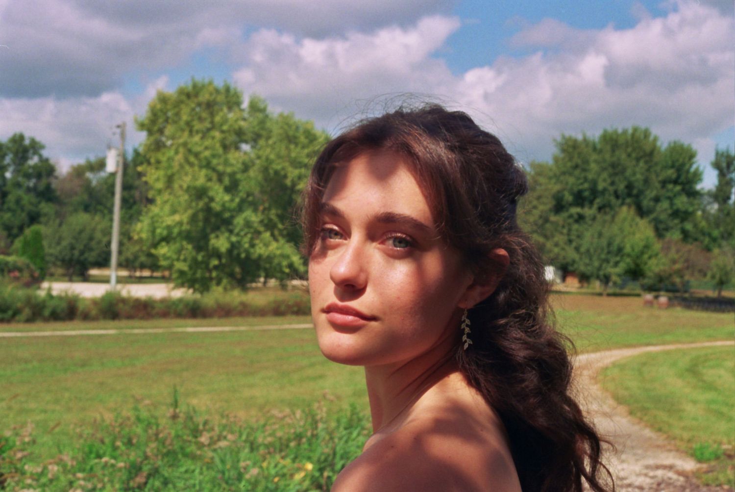 A person with long dark hair poses thoughtfully in a sunny field with green trees and cloudy skies in the background.