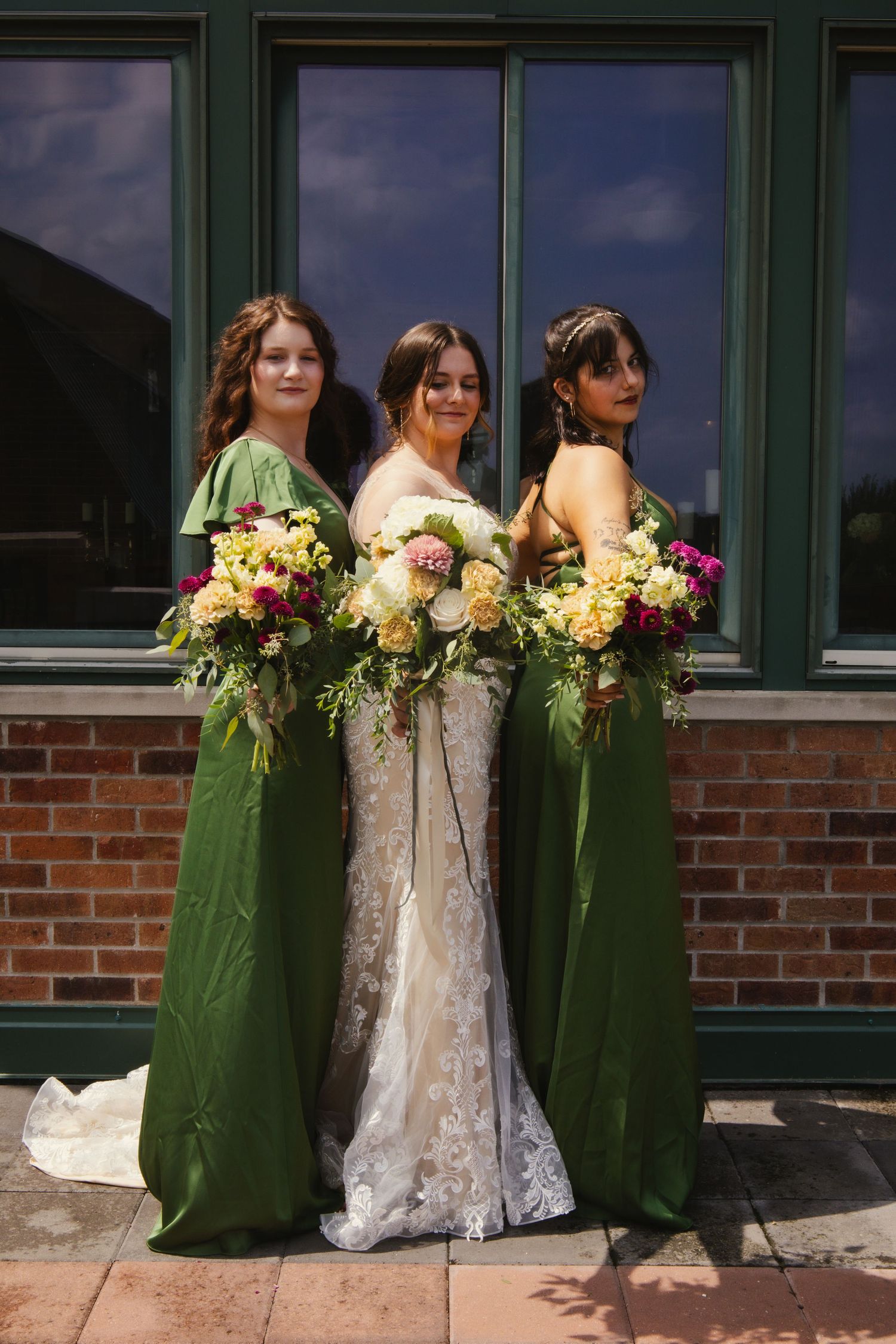 Three bridesmaids in elegant green dresses and a bride in white lace gown holding colorful floral bouquets pose outdoors.