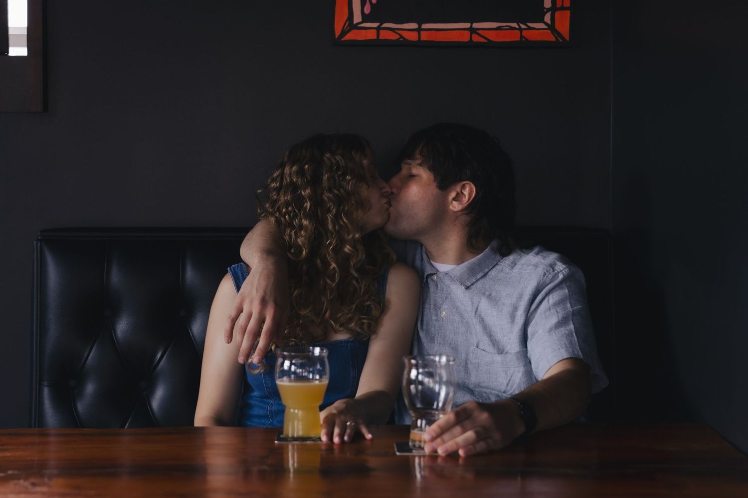Couple kissing romantically at a bar table with beer glasses, seated on a black leather booth.